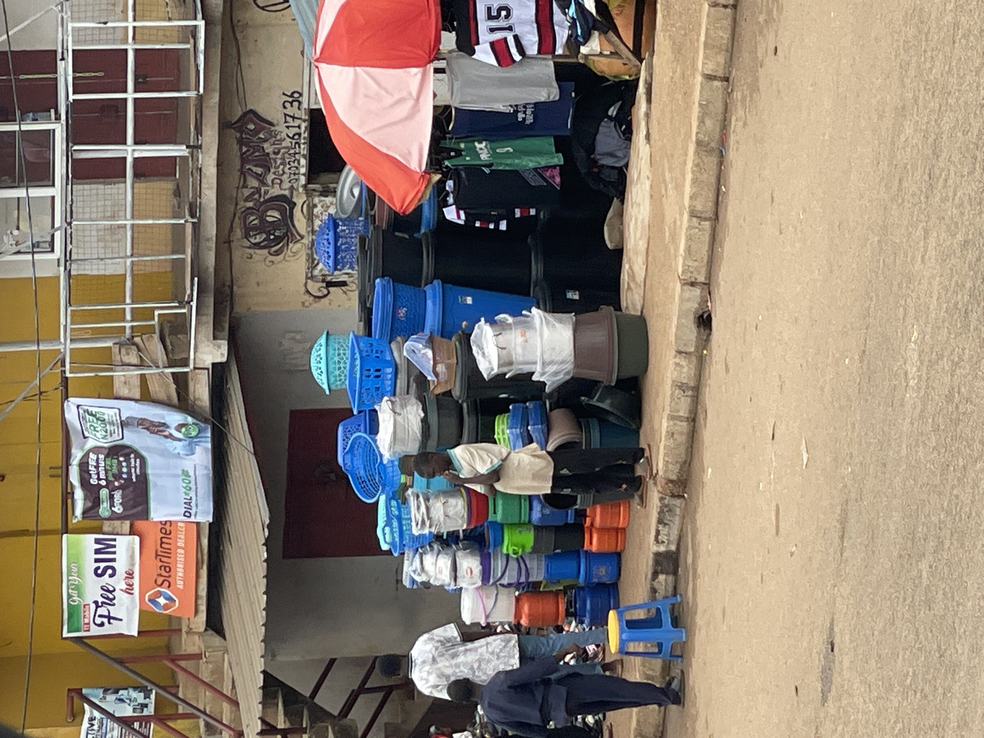 The image shows a street scene in Bukuru, Nigeria. A vendor has set up a shop on the sidewalk, selling a variety of goods, including clothing, baskets, and buckets. The merchandise is stacked high. A red and white umbrella provides shade. Two people are visible near the vendor's display. The street is paved. Buildings with signs are visible in the background. Signs for "Free SIM" and "StarTimes" can be seen. Graffiti is present on the building wall.
