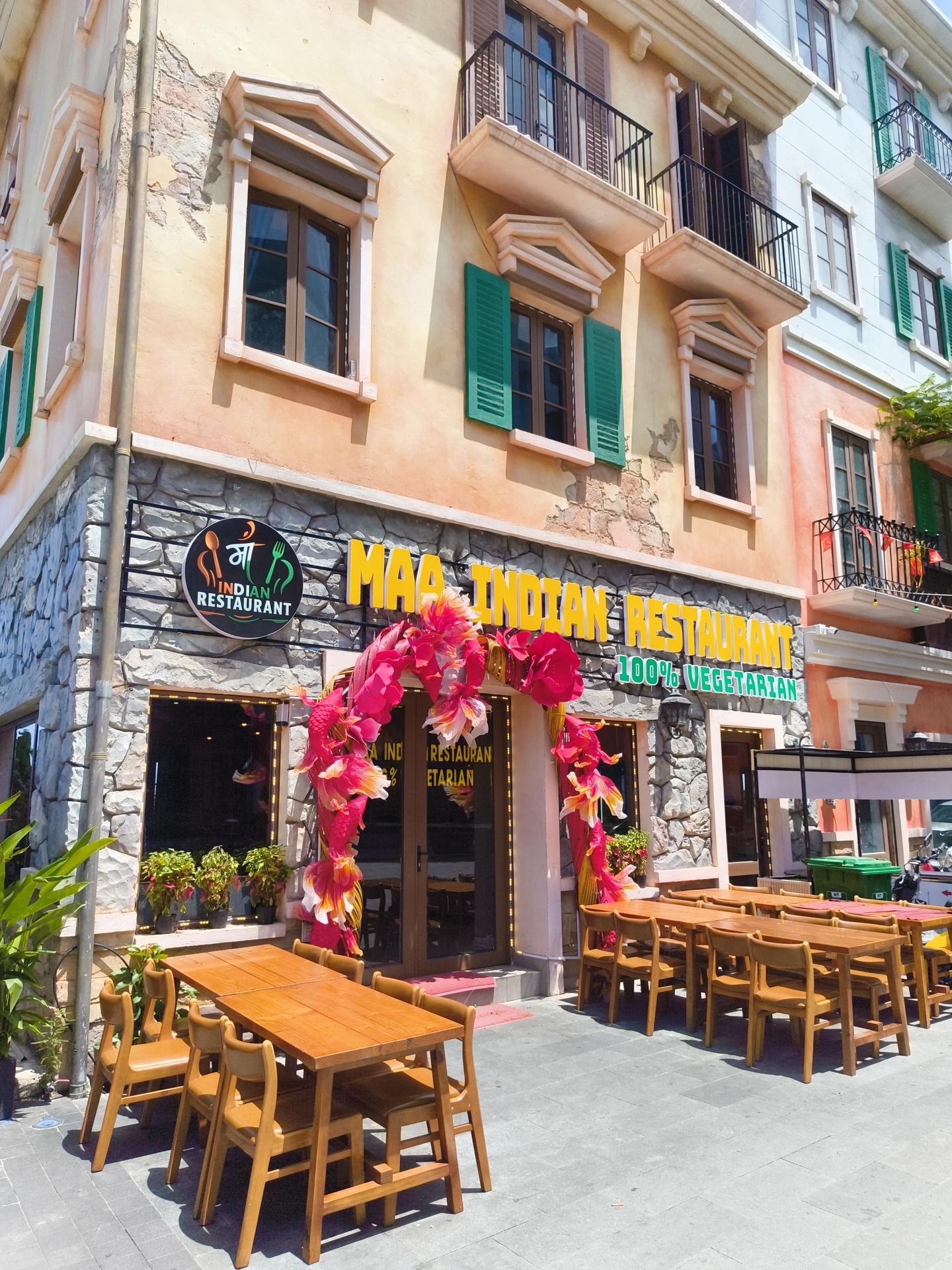 This is a medium shot, taken outdoors, of the exterior of a restaurant in Phu Quoc, Vietnam. The building facade is a mix of faux stone and stucco, painted in warm tones of orange and yellow. The architecture features decorative window frames and balconies. In front of the restaurant is an outdoor seating area with wooden tables and chairs. A prominent sign above the entrance reads "MAA INDIAN RESTAURANT 100% VEGETARIAN" in large yellow letters. The entrance is adorned with a colorful floral archway, primarily in shades of pink and yellow. The time of day appears to be daytime, with bright sunlight casting shadows. No people are visible in the frame.