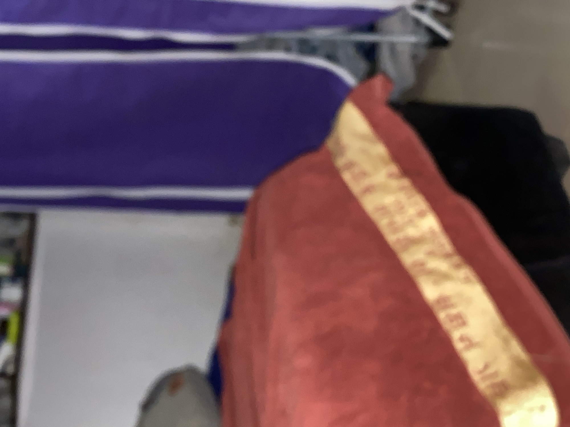 The image depicts a close-up, unfocused view of a bed covered with a red blanket adorned with a gold trim. The gold trim has some visible text or pattern on it, though it is blurry. In the background, a purple curtain with white vertical stripes is visible, partially obscuring some objects. To the left of the bed, a grey pillow or cushion is partially visible. The overall environment appears to be indoors, possibly a bedroom. The image does not provide any specific clues about its location in Jalingo, Nigeria.