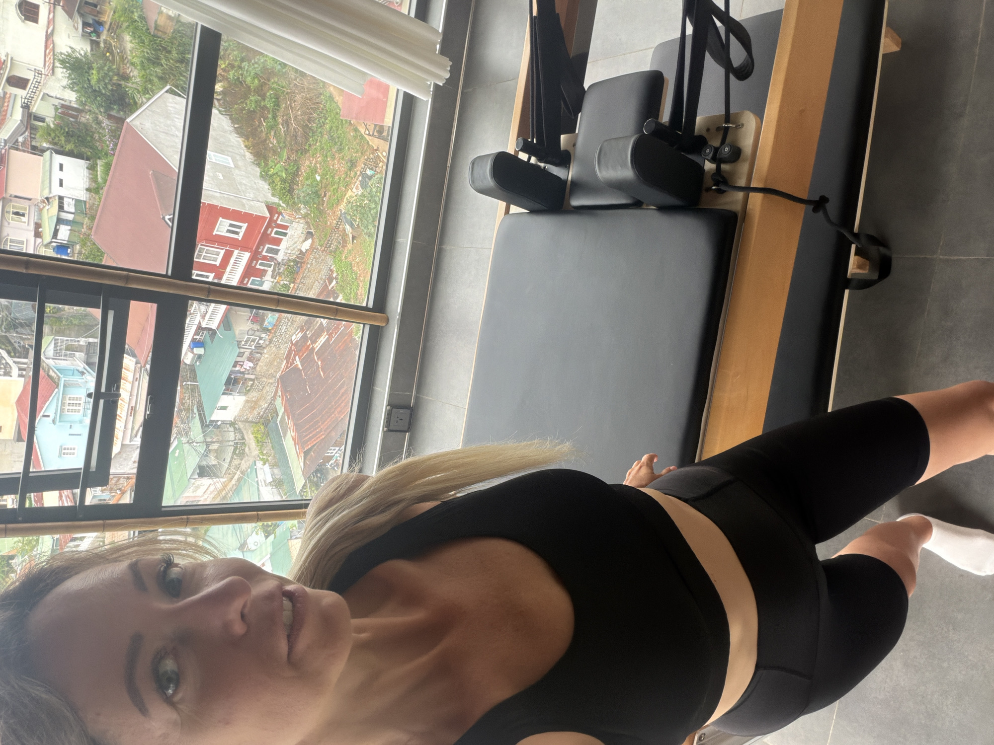 A blonde woman in her 30s, wearing a black sports bra and black bike shorts, poses for a selfie in front of a Pilates reformer. She stands in what appears to be an apartment with a large window overlooking a densely populated hillside city. The city has a mix of building styles and colors, with many red-tiled roofs. In the foreground, the dark grey floor is visible, as is a white sock on the woman's left foot. The woman's hair is long and blonde, pulled back from her face. The Pilates reformer is made of wood and black upholstery, with various straps and attachments. The background shows a vibrant urban landscape, characteristic of Da Lat, Vietnam.