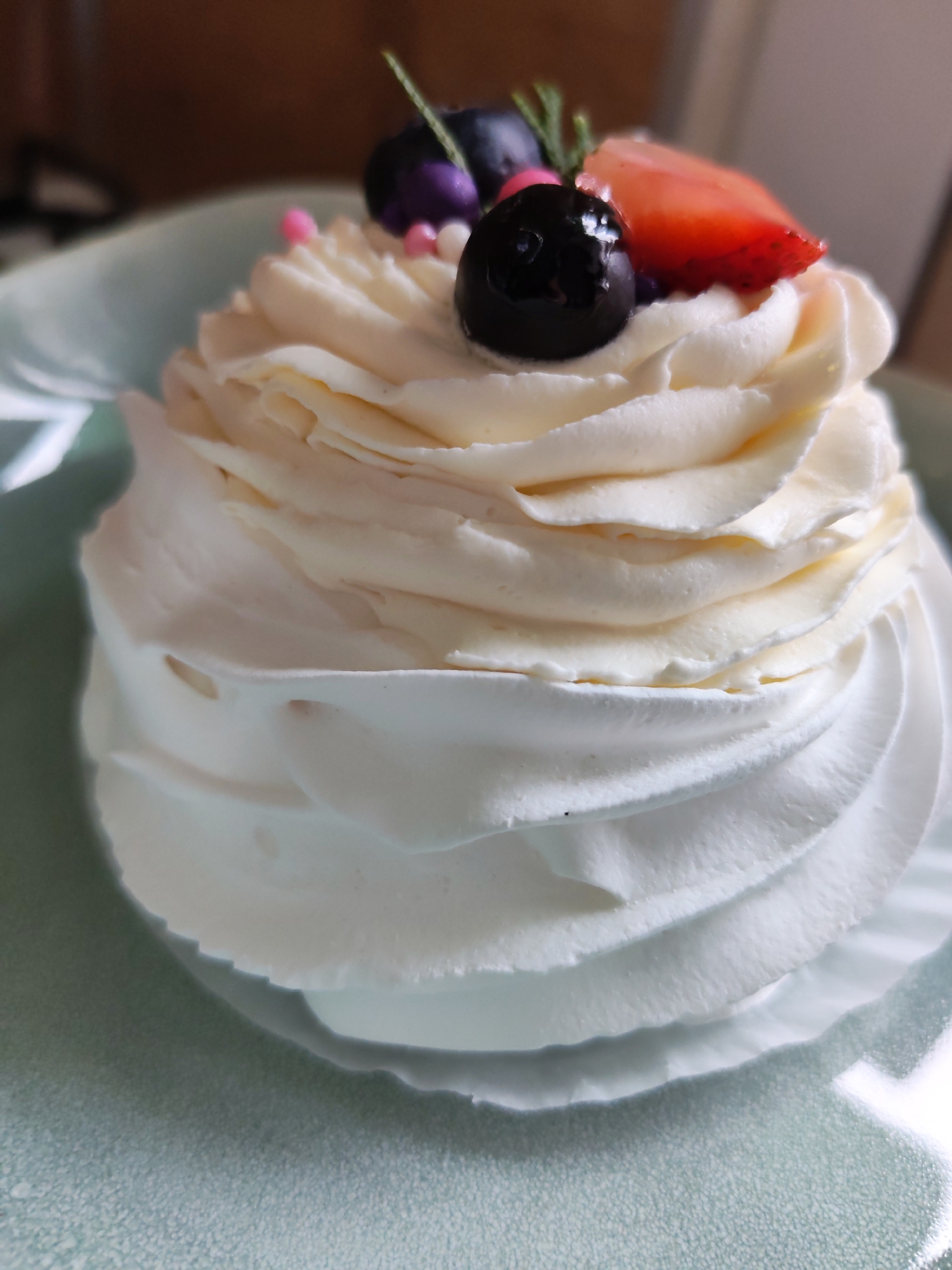A close-up shot captures a Pavlova dessert on a pale green plate. The dessert is constructed from a meringue base with swirls of cream, topped with fresh blueberries and a slice of strawberry. It is sitting on a plate on a table in what seems like a home environment. There are no identifiable people or specific actions. The location of Izhevsk, Russia is not discernible from the image.