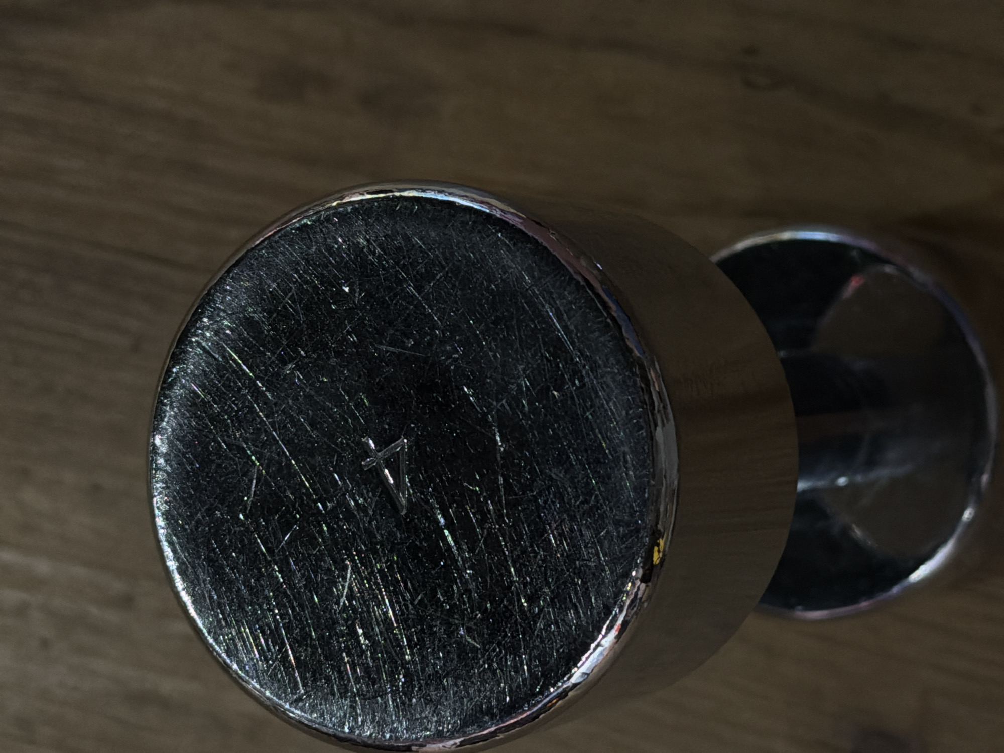 A close-up, top-down shot of a scratched metal dumbbell on a wooden surface. The dumbbell's top plate, likely chrome-plated, exhibits numerous fine scratches, reflecting light in iridescent hues. The wooden surface appears to be floorboards. The scene is devoid of any human presence or activity. The image does not provide any information to locate it within Strasbourg, France.
