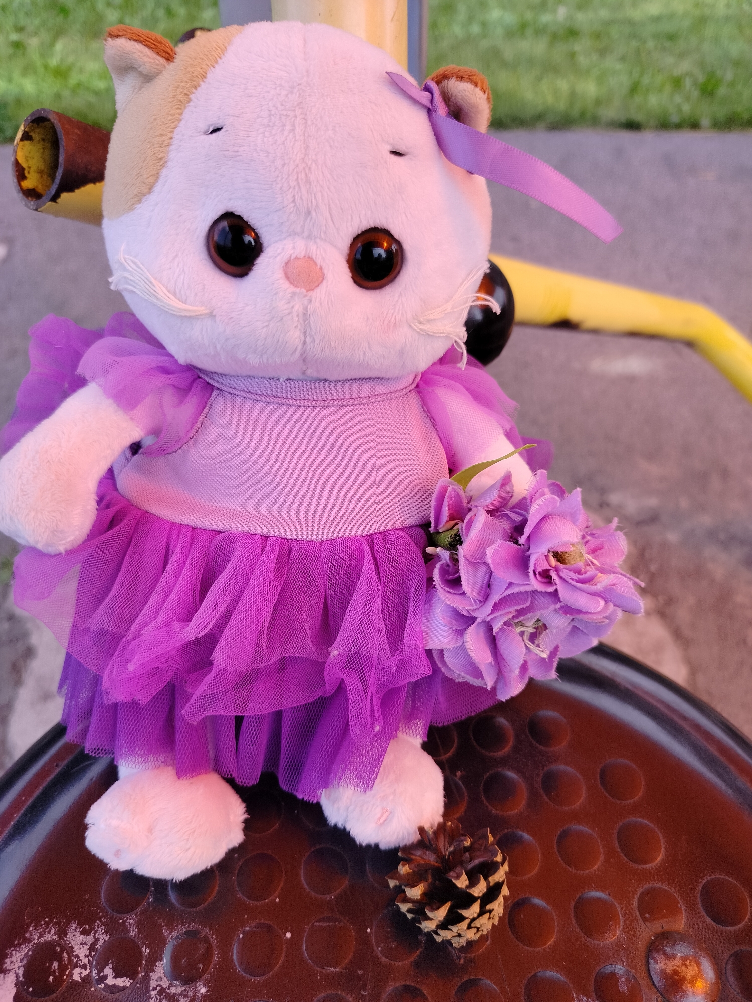 A stuffed cat wearing a purple tutu and holding a bouquet of purple flowers sits on a dark brown textured surface. A small pinecone is on the surface next to the cat. The background is blurred green grass and yellow and brown playground equipment. The image was taken in Yashkino, Russia.