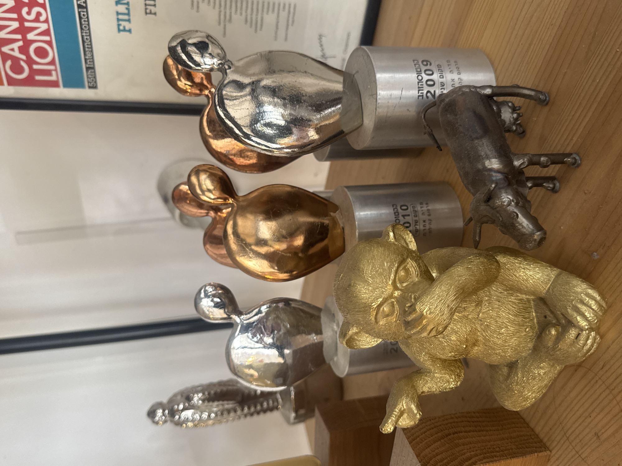 The image shows a collection of awards on a wooden surface. There are three awards with rounded, bulbous shapes in different metallic finishes: silver, bronze, and silver again. Each is mounted on a cylindrical base, which are labelled in Hebrew, with the years "2009" and "2010" visible. These awards appear to be Cannes Lions awards, according to the text in the upper left corner of the image. There are two other statuettes, one of an animal, a pig, made from welded metal, and another one of a golden monkey. The image was taken in Tel Aviv, Israel.