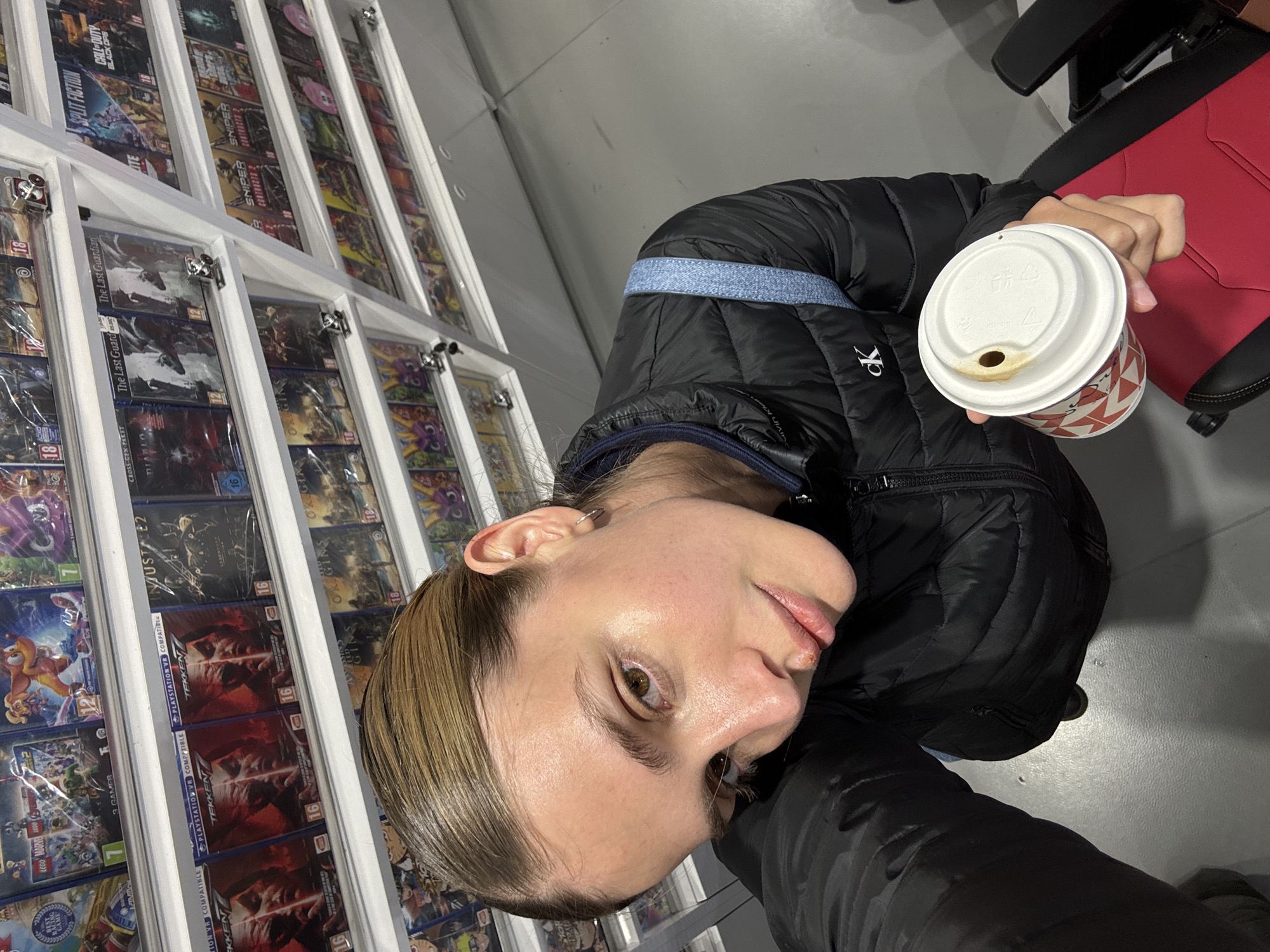 A person stands next to a shelf filled with video game boxes while holding a cup of coffee. The titles visible on the game boxes include "LEGO Marvel," "Tekken," "Injustice 2," "The Last Guardian," "Split Fiction," "Diablo," "Origins," and "Call of Duty Black Ops." The person is wearing a black jacket. The scene takes place in a store, likely a video game retailer. The location is Tbilisi, Georgia.