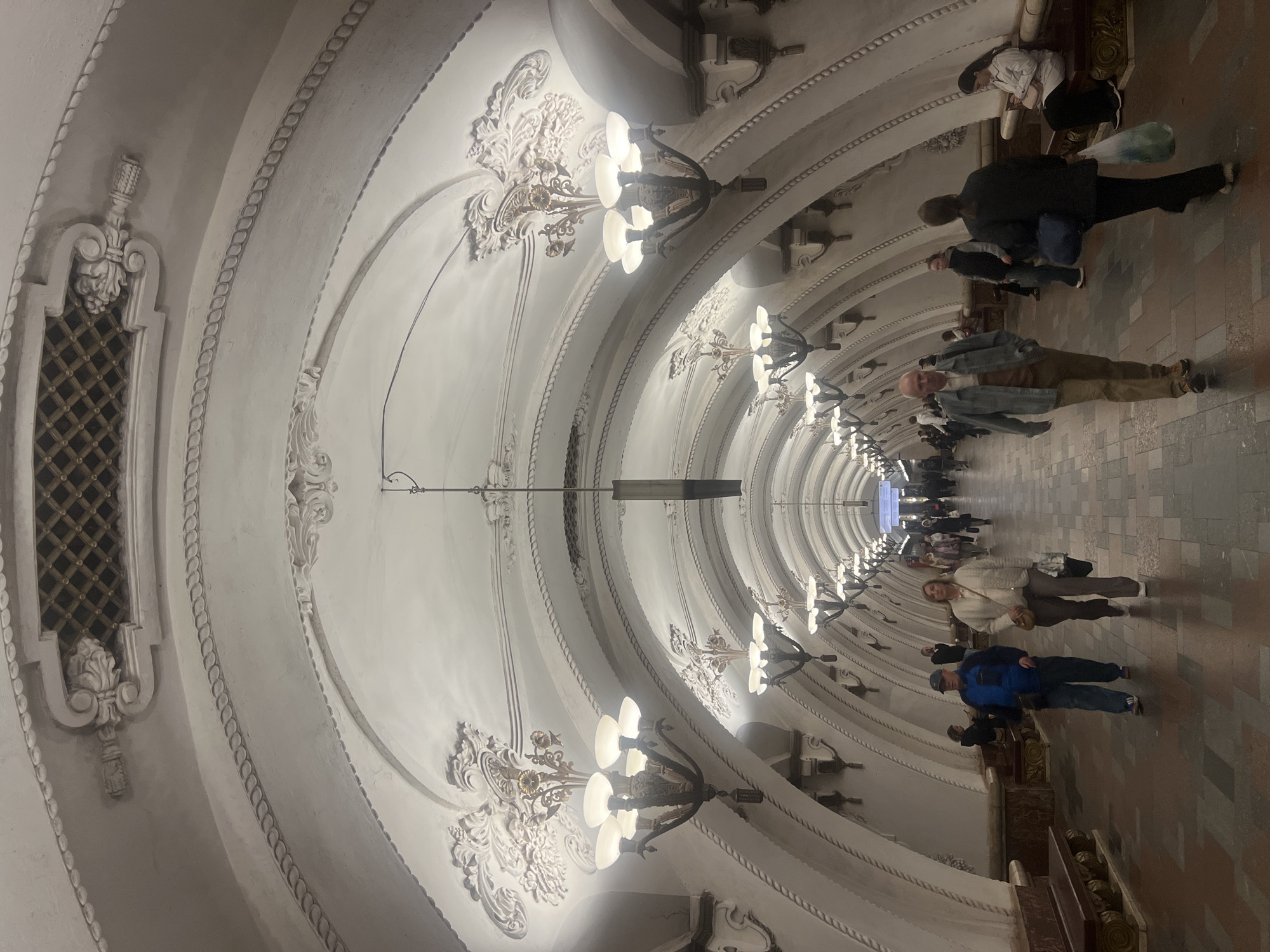 The image shows an interior view of a long hallway or tunnel. The ceiling is arched and ornate, decorated with intricate white plasterwork, including rows of chandeliers. The floor is tiled in a gray-brown pattern. Several people are walking along the hallway, appearing as blurred figures. The tunnel stretches far into the distance, narrowing to a vanishing point. The location appears to be a subway station.