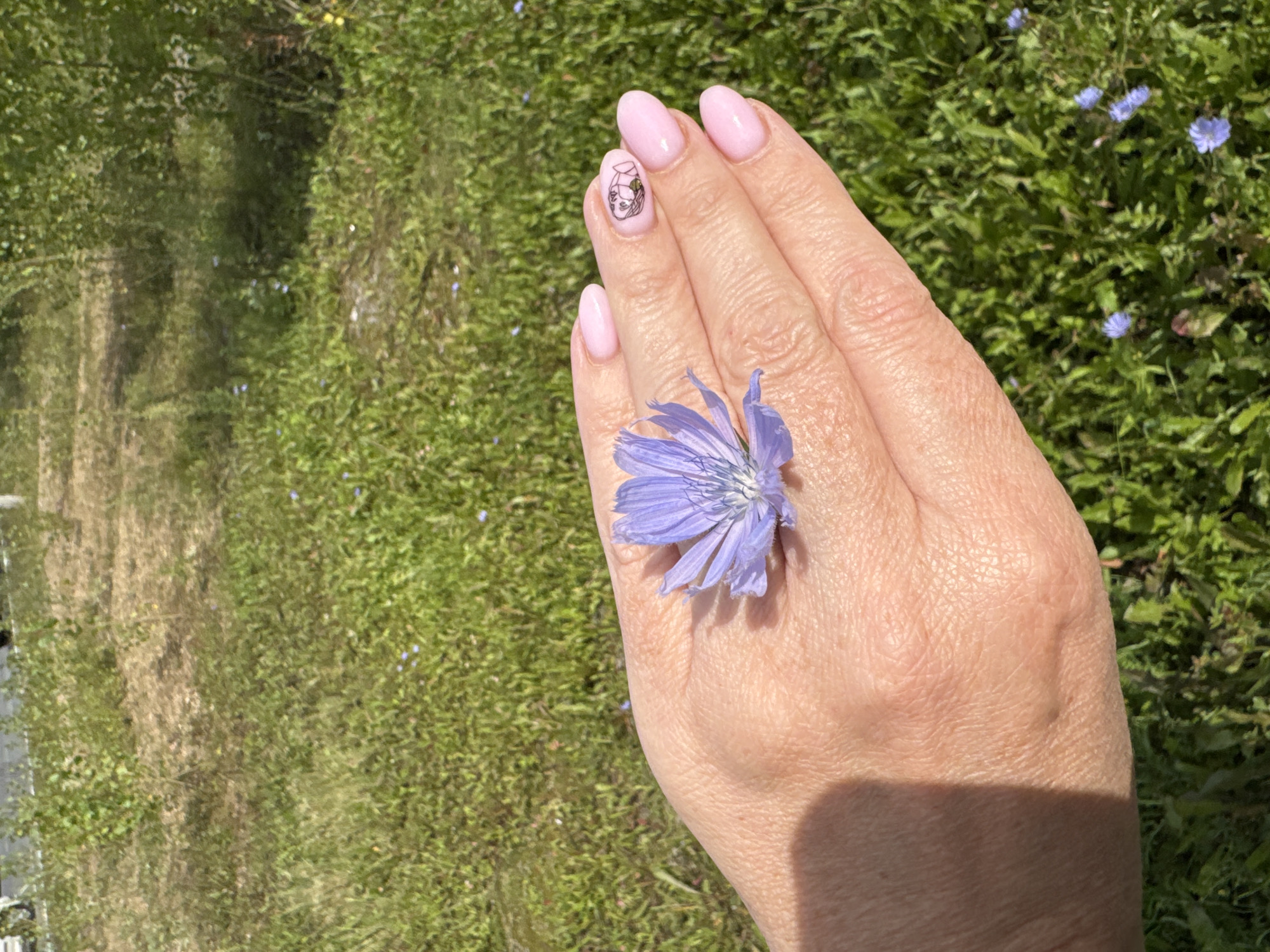 A person's hand, with light pink painted fingernails, one of which has a small black drawing of a face, is held open palm up. A single purple flower with delicate petals is placed on the palm. The background is a grassy area with scattered small blue flowers, suggesting a natural outdoor setting. The image does not provide enough information to determine the specific location within Ufa, Russia.