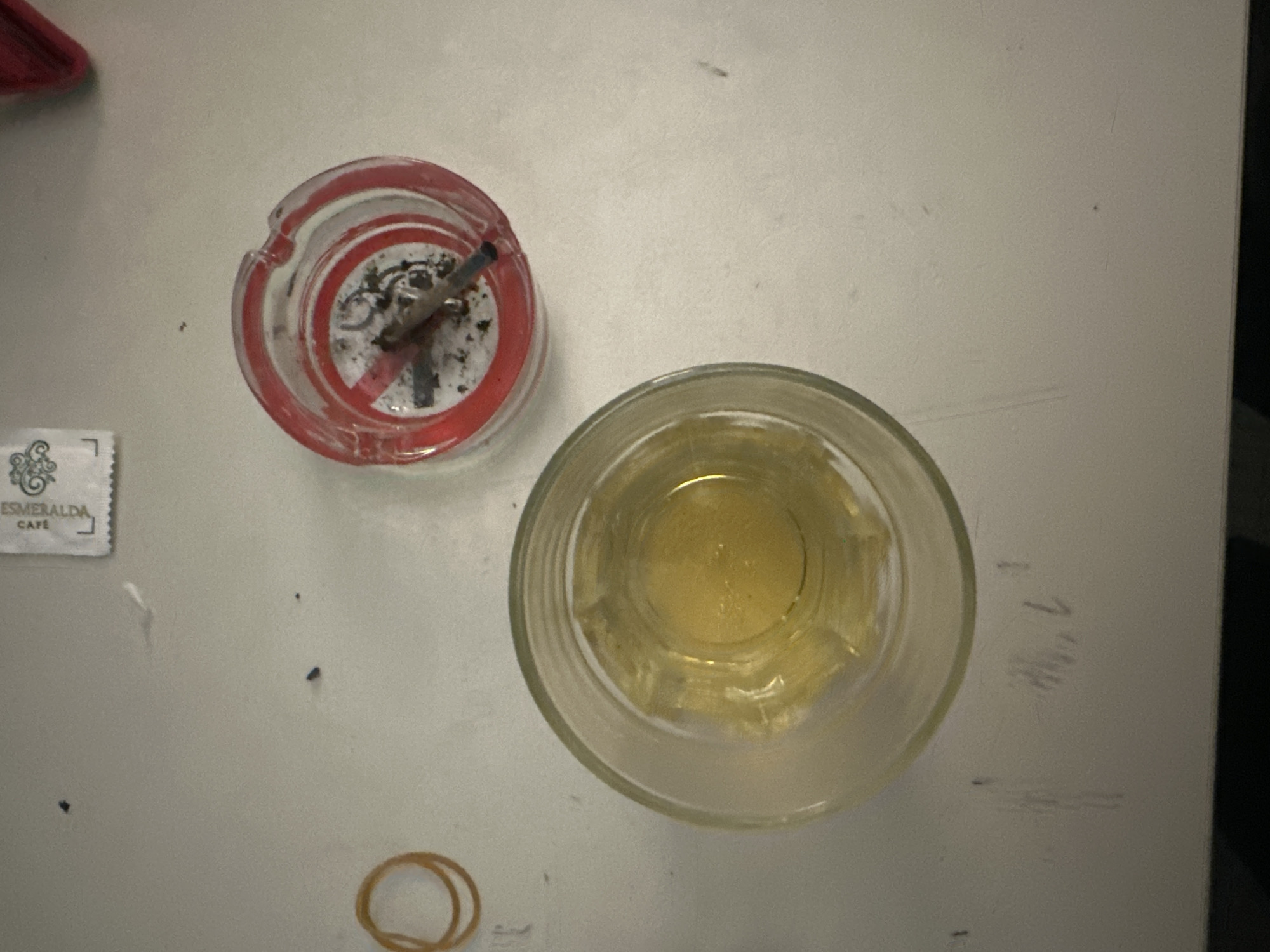 The image displays an overhead shot of various items resting on a light-colored, possibly white, surface that appears somewhat scuffed or marked.
**Subjects:**
The primary subjects are a glass ashtray, a drinking glass, a small packet, and a rubber band. The ashtray is made of clear glass with red detailing, specifically a prominent "no smoking" symbol (a red circle with a diagonal line through it) which appears to be crossed out or defaced. It contains the butt of a cigarette and ash. Adjacent to the ashtray, a clear drinking glass holds a yellowish liquid, possibly an alcoholic beverage like whiskey or a light-colored soft drink. In the bottom left corner, a small white rectangular packet is visible, and near it, a brown rubber band. A small, blurry red object is partially visible in the top left corner of the frame.
**Activity or event taking place:**
No individuals are present, but the arrangement of items suggests a recent or ongoing moment of relaxation or leisure, possibly involving smoking and drinking. The scene could be set at a bar, cafe, or a personal space.
**Notable details:**
The time of day appears to be evening or night, or at least in a dimly lit indoor setting, given the overall lighting in the image. The surface beneath the objects shows wear and tear. A particularly notable detail is the "no smoking" symbol on the ashtray itself, which is ironically being used to hold a cigarette. The liquid in the glass has a warm, inviting hue.
**Visible text:**
The text "ESMERALDA CAFÉ" is clearly visible on the small white packet in the bottom left, alongside a decorative swirl.