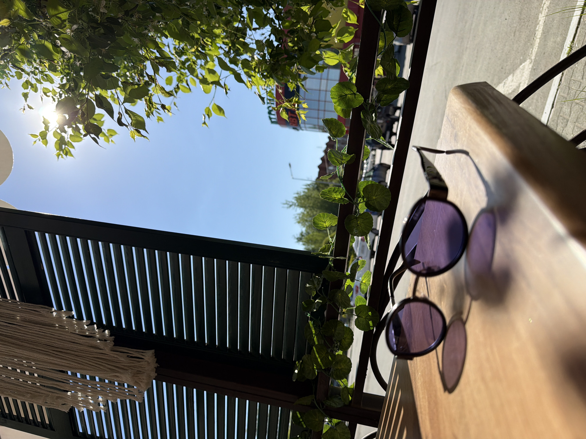 The image shows a sunny outdoor scene likely taken from a cafe or similar establishment. In the upper left, the sun peeks through tree branches with bright green leaves. The sky is a clear blue.
A dark-colored, slatted structure is partially visible, with a white macrame hanging from it on the left. In the middle ground, artificial vines are draped along a railing. In the distance, a building with a red and green sign is visible.
A pair of round, purple-tinted sunglasses rests on a light-colored wooden table in the foreground. The table casts a sharp shadow. The image seems to be taken on the outdoor area of a restaurant or cafe. Based on the prompt mentioning Chaikovsky, Russia, one could assume the photo was taken in that city.