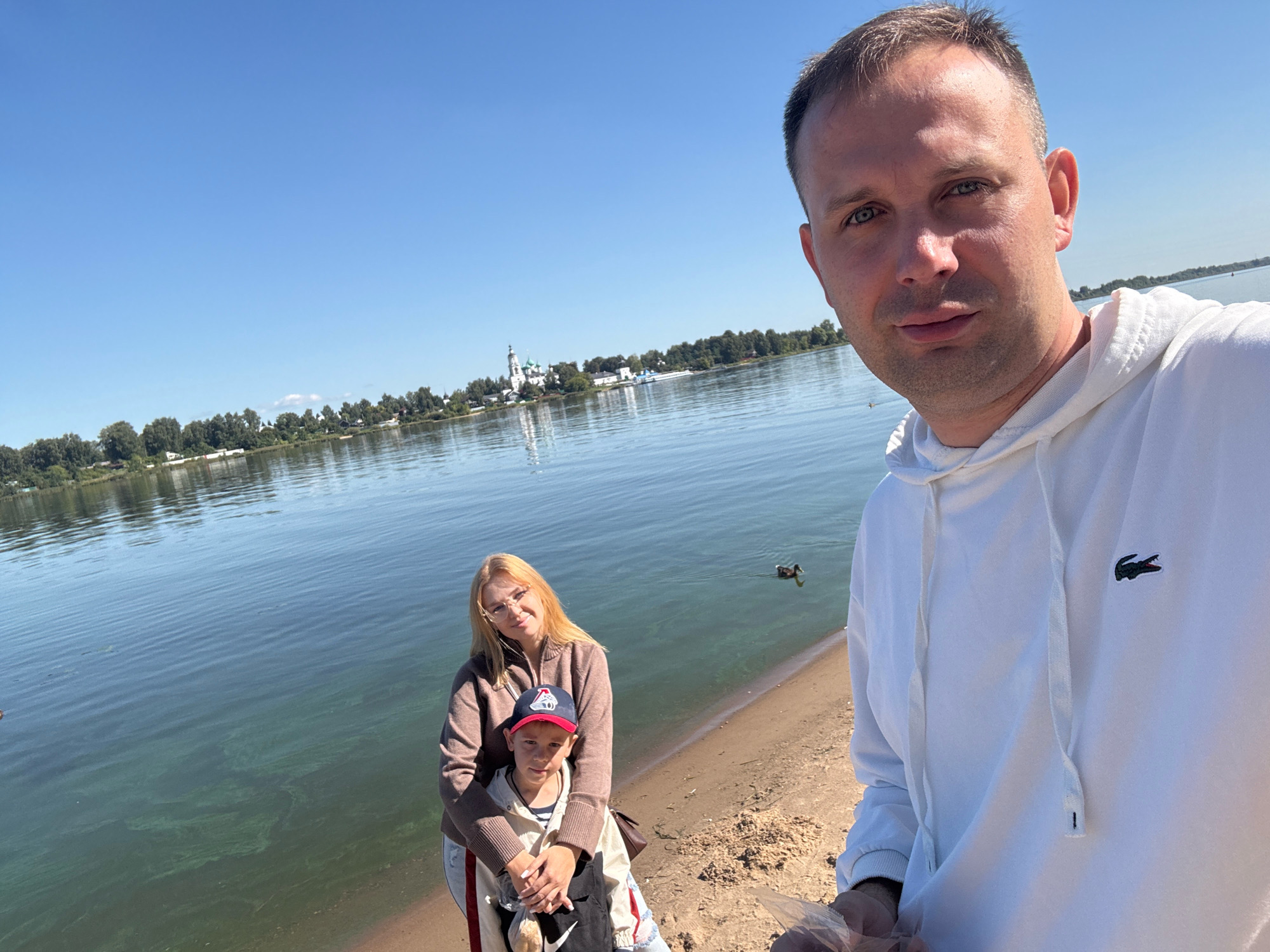 In Ignatovo, Russia, a group of people is pictured at a waterfront on a sunny day. A man in a white hooded sweatshirt takes a selfie, with his face filling the right side of the frame. Behind him, a woman in a brown cardigan and a young boy in a cap stand on a sandy shore. The water is calm and reflects the clear blue sky. In the distance, a white building with green domes, likely a church, is visible on the far bank, surrounded by trees. A duck swims in the water near the shore. The overall scene depicts a pleasant outdoor excursion by the water.