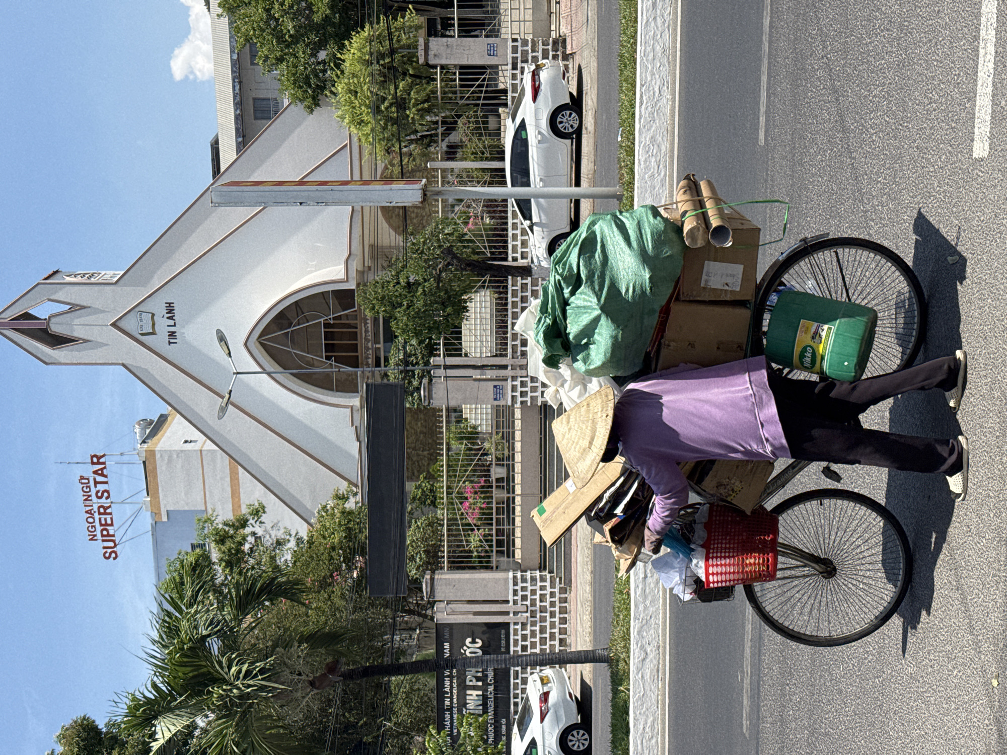 A person wearing a purple shirt and a traditional Vietnamese conical hat is riding a bicycle loaded with various items on a paved road. The bicycle has a red basket in the front and a green bag, cardboard boxes, and other miscellaneous items piled on the back. The person is wearing black pants and white sneakers.
In the background, there is a church-like building with the words "TIN LANH" inscribed above the entrance. There are also trees, buildings, and a sign that reads "NGOAI NGU SUPER STAR". There is also a building named, "Hội Thánh Tin lành Việt Nam." The scene is set in a city, presumably Nha Trang Ward, Vietnam. A couple of white vehicles are seen parked next to the road. The weather appears to be sunny.
