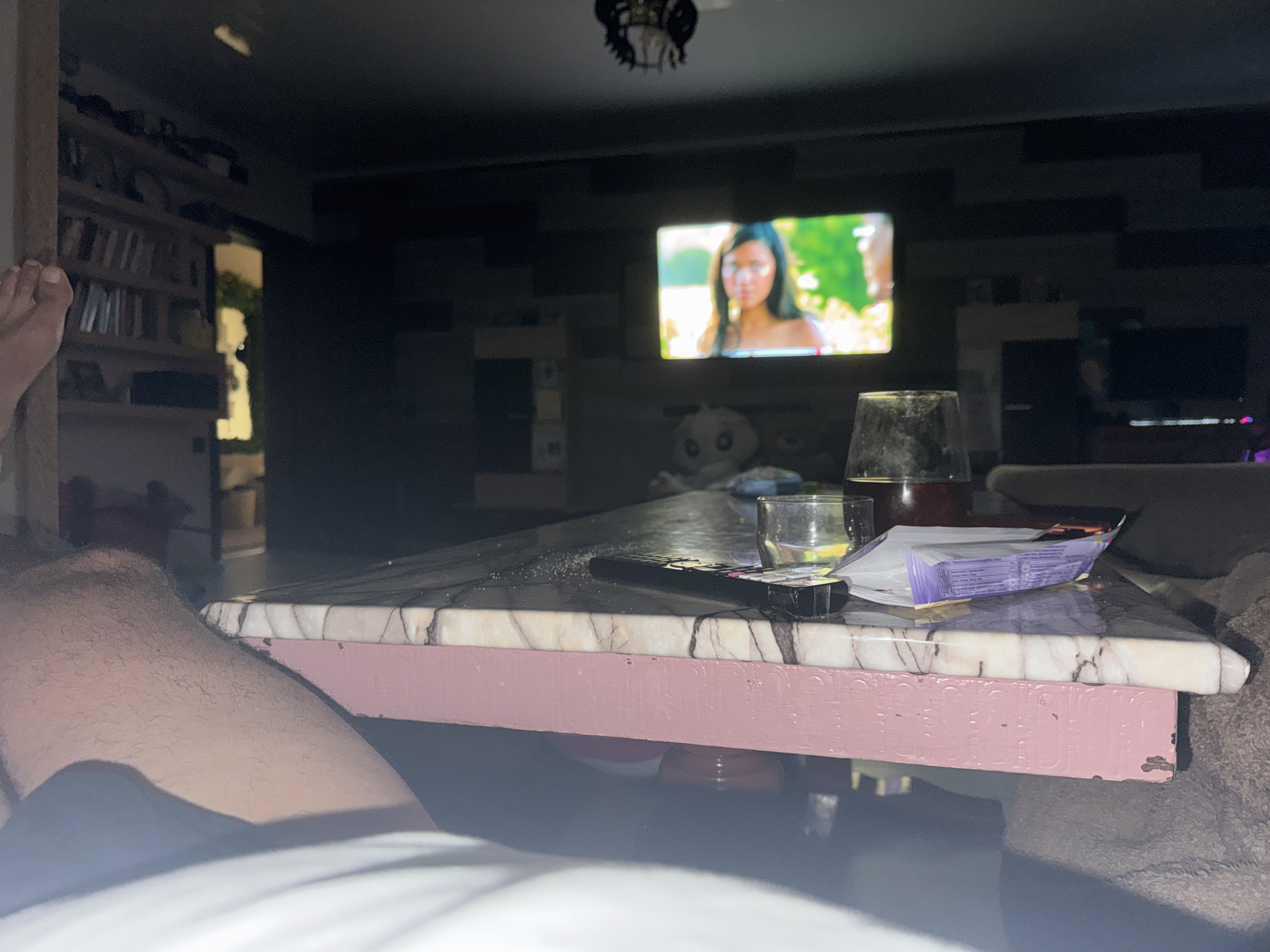 This image depicts an indoor scene, likely a living room, with a low-angle perspective. In the foreground, a person's leg and foot are visible, suggesting they are reclining. A table with a marble-patterned top and a pink base occupies the center of the frame. On the table, there is a remote control, a small glass with a yellow liquid, a larger glass with a dark liquid, and a partially unwrapped package of what appears to be candy or snacks. In the background, a television screen displays a woman's face. Shelves with books and various objects are visible on the left, and a dark entertainment unit with a television is on the right. The lighting is dim, with the television screen being a primary light source. The overall impression is one of a relaxed, domestic setting. The location in Svilengrad, Bulgaria is not determinable from the visual information provided in the image.