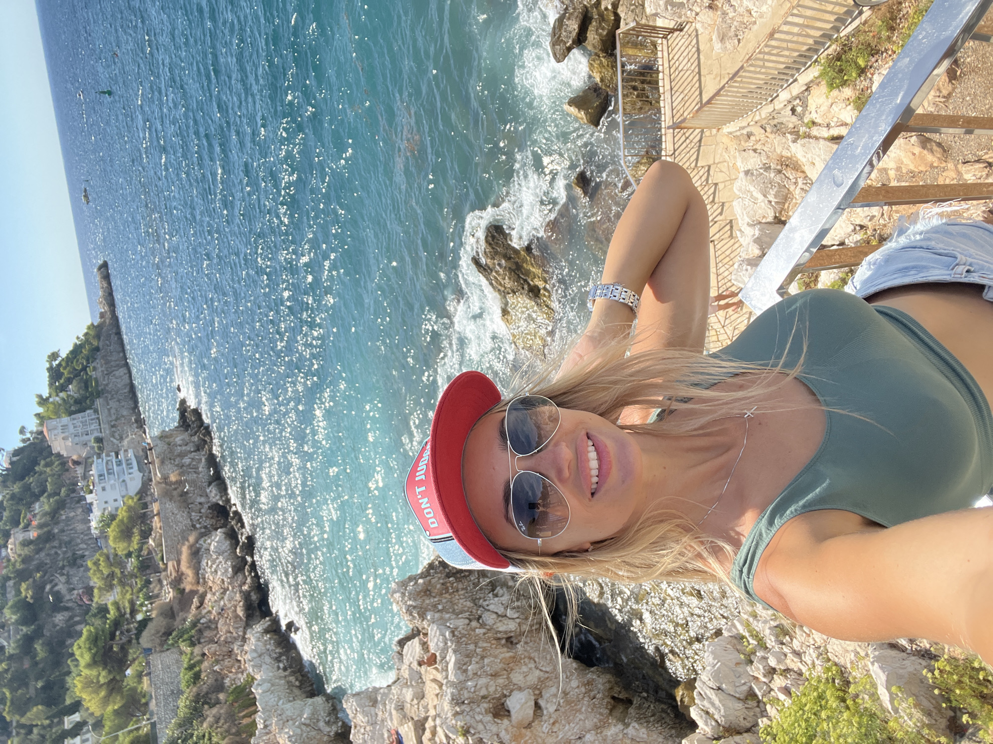 In the city of Nice, France, a blonde woman with a red and blue baseball cap and aviator sunglasses is taking a selfie on a sunny day. She is wearing a green tank top and denim shorts. Behind her, the turquoise water of the Mediterranean Sea crashes against rocky shores, and a stone pathway with a metal railing is visible. In the distance, white buildings are nestled on a green, tree-covered hillside.
