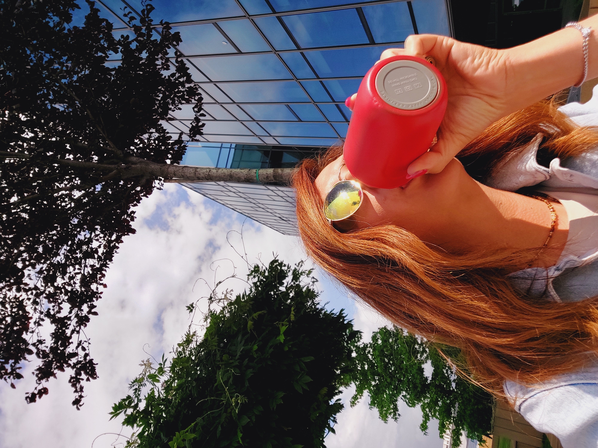 A woman with long, reddish-brown hair, wearing round sunglasses and a denim jacket, is seen from the chest up. She is holding a bright red reusable water bottle to her lips, as if drinking. The background features a modern building with blue glass windows, a tree with dark leaves partially obscuring the sky, and a cloudy sky. The scene appears to be outdoors, possibly in an urban park or plaza in Sofia, Bulgaria.