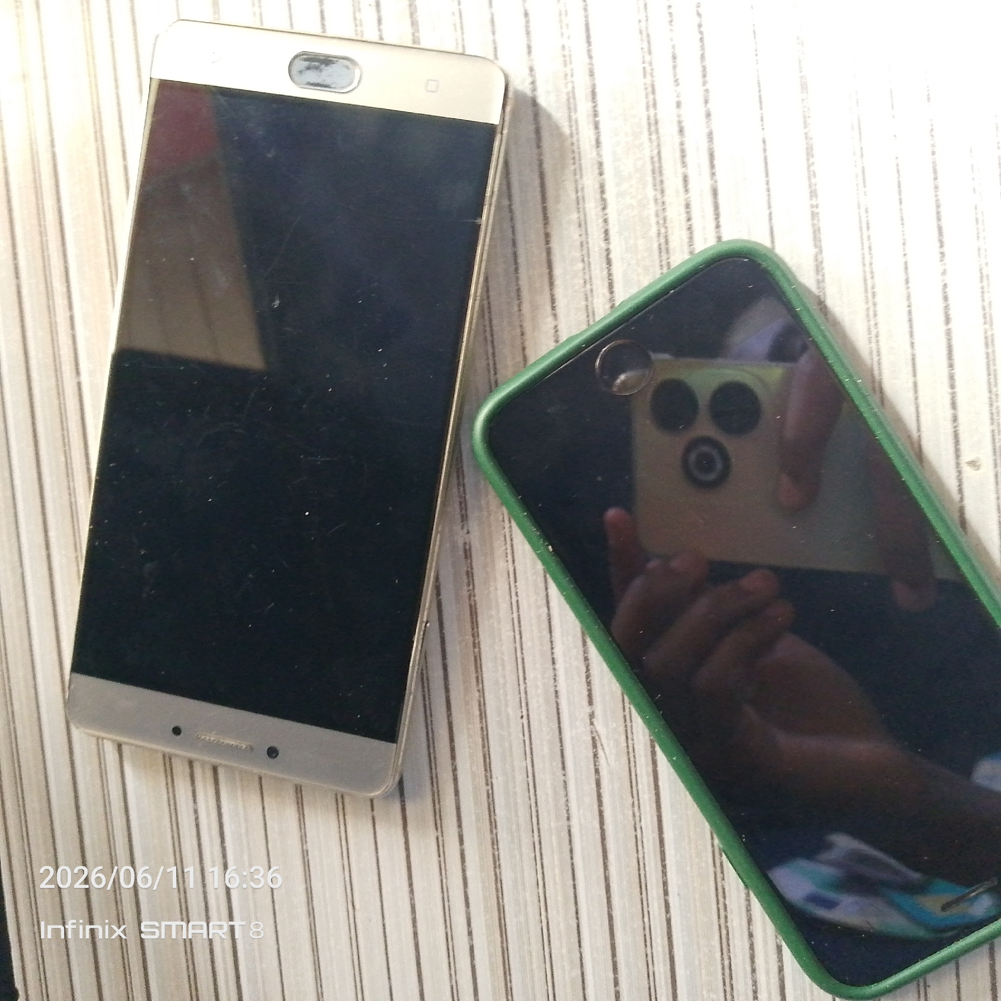 Two smartphones are placed on a light-colored, vertically striped surface. The phone on the left is gold-colored with a large black screen. It has a silver element at the top with what appears to be a speaker or camera cutout. The bottom edge of this phone has two small black dots and a metallic strip. The phone on the right has a dark screen and is encased in a bright green cover. Its camera module, visible in the reflection on the screen, is triangular with three circular lenses and a smaller circular element in the center. The reflection also shows a person's hands holding the phone and a partial reflection of another device with a yellow casing and circular lenses. A watermark in the bottom left corner of the image reads "2026/06/11 16:36 Infinix SMART 8". The scene is indoors, and the lighting suggests it is daytime, with soft shadows cast by the phones.