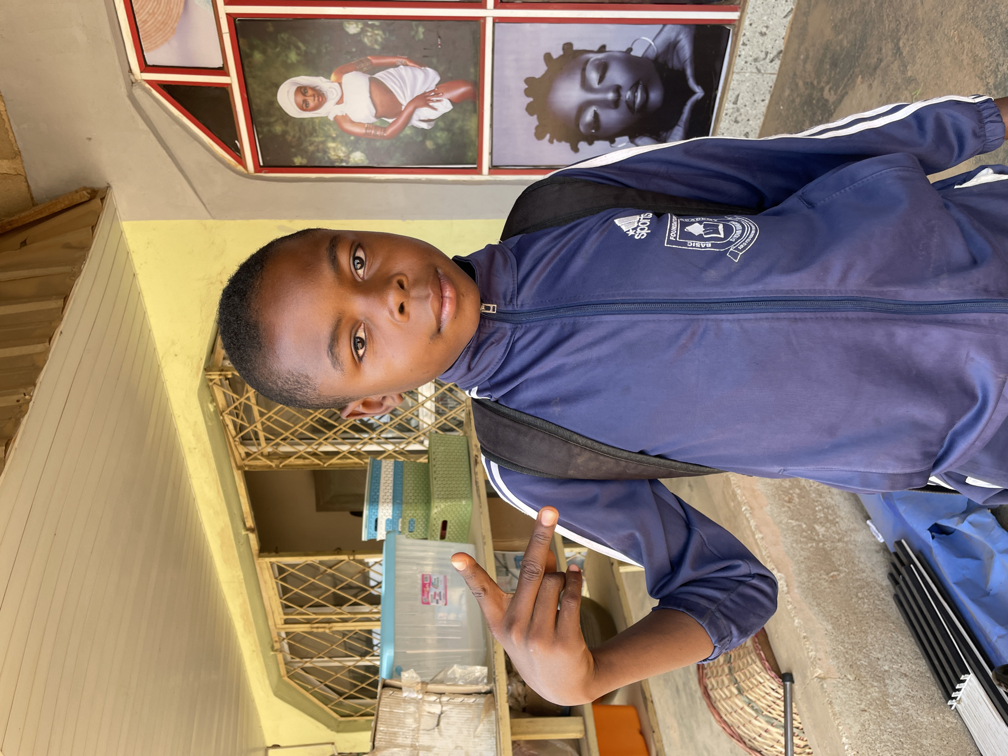 A young Black male, appearing to be a teenager, is the central subject of this portrait. He looks directly at the camera with a calm, neutral expression. He is wearing a dark blue tracksuit jacket with white stripes on the sleeves, characteristic of Adidas, and a black backpack. On the left chest area of his jacket, the word "sports" is visible above the Adidas logo, and below it, a circular emblem partially shows text including "FOUNDATION" and "BASIC". His right hand is raised, making a gesture where his thumb and pinky finger are extended, and his middle fingers are curled.
The setting appears to be outdoors or within a semi-open structure in Barakin Akawo, Nigeria, during the daytime. Behind the subject, a light yellow exterior wall is visible, with architectural details including a window or opening secured with metal grates. Above and to the left of the subject, a section of the wall features four framed images. Two are clearly discernible: one showing a person with dark skin, dressed in white fabric and a headwrap with bangles, against a green background; and another in black and white, portraying a person with dark skin and dreadlocks, with closed eyes.
Further into the background, various objects are stacked and arranged, including several light blue plastic storage bins or containers and what appear to be cardboard boxes. On the ground to the right, a woven basket and several metal rods or poles are visible, along with a partially obscured blue tarp. The lighting is bright and even, indicating it is daytime. The overall environment suggests a casual, possibly commercial or residential, outdoor space.
