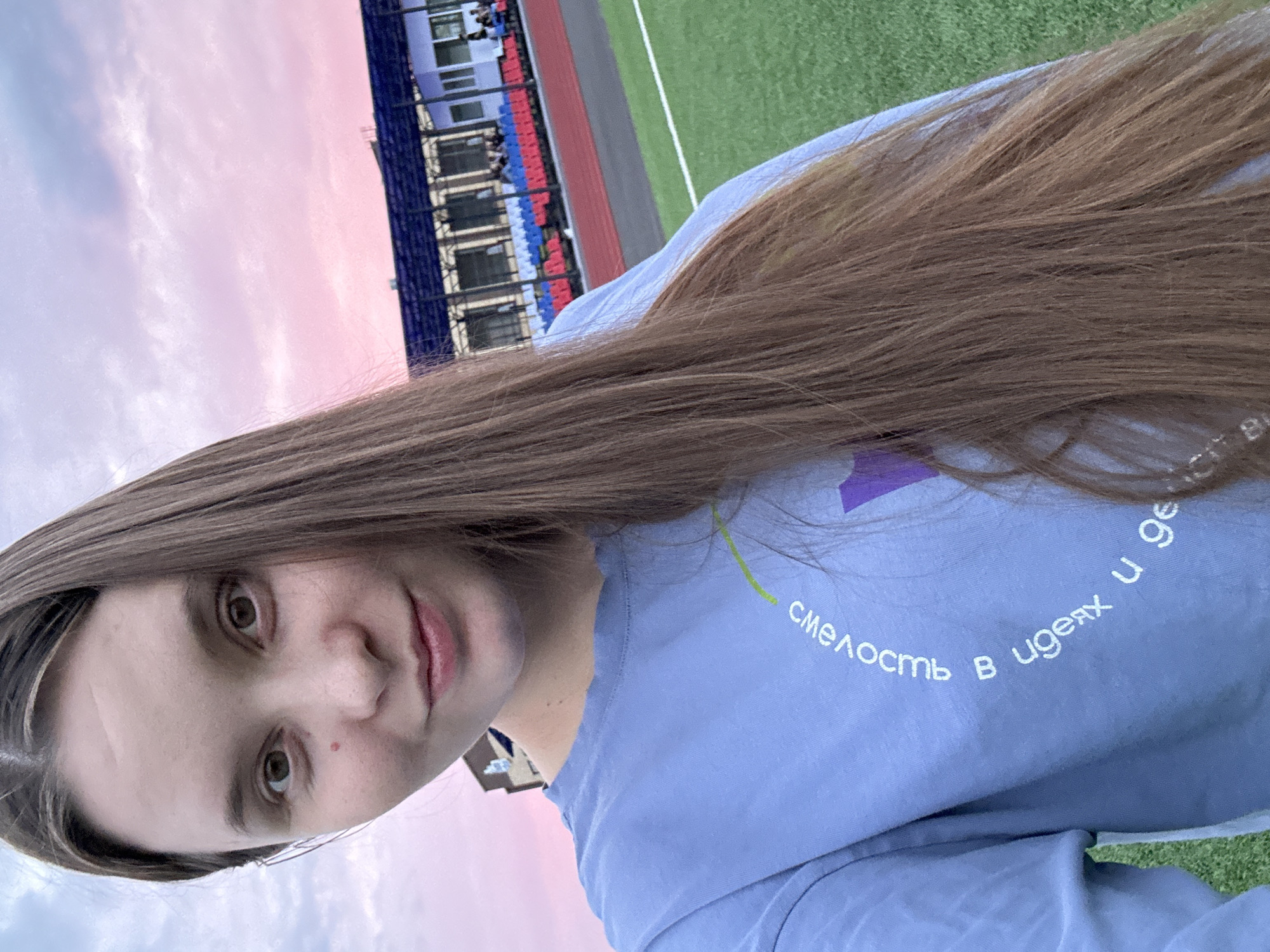A young woman with long brown hair takes a selfie outdoors in front of a stadium with a pinkish-purple sunset sky. She is wearing a light blue long-sleeved shirt with white Cyrillic text and a purple graphic on it. The stadium has blue and red seating and a green sports field. The image appears to be taken during daylight with the sky transitioning to dusk.
