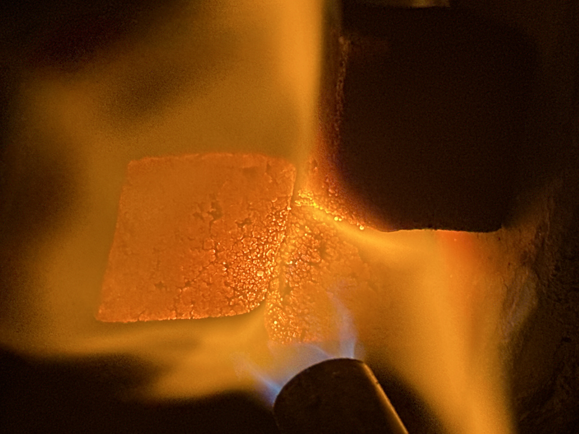 A close-up shot depicts a soldering torch heating an object. The object, which appears to be a cube with a textured surface, is glowing orange from the heat. The torch nozzle, visible in the lower portion of the image, emits a blue-tinted flame. The surrounding environment is dark, providing contrast to the bright orange glow.