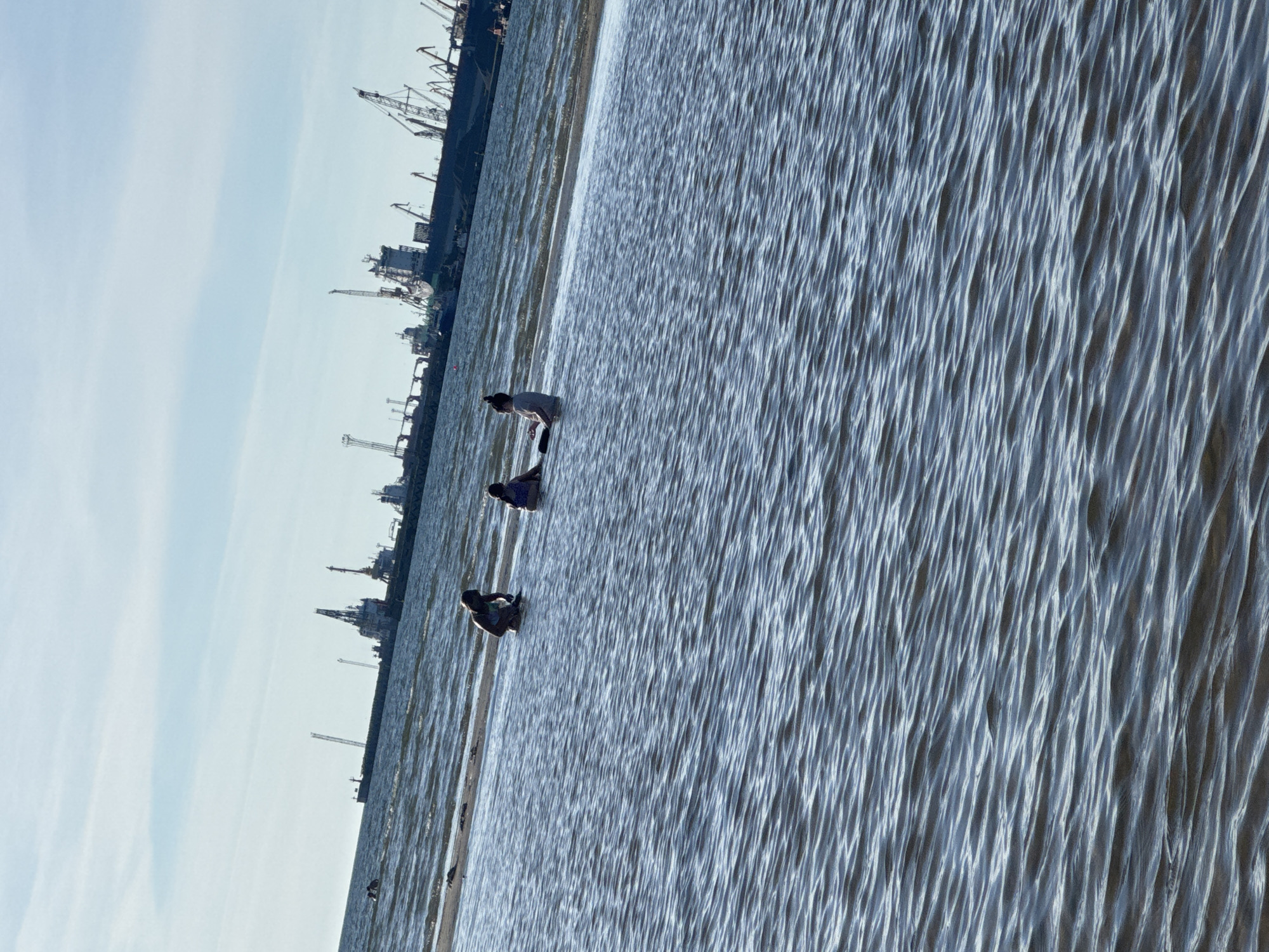 Three individuals are partially submerged in shallow water, with a sandy area visible behind them. In the background, a large cargo ship with numerous cranes is docked at a port. The sky is clear with a few wispy clouds, and the water's surface is rippled. This scene is likely located in the port area of Tallinn, Estonia, potentially near the Tallinn Passenger Terminal or the Old City Harbor.
