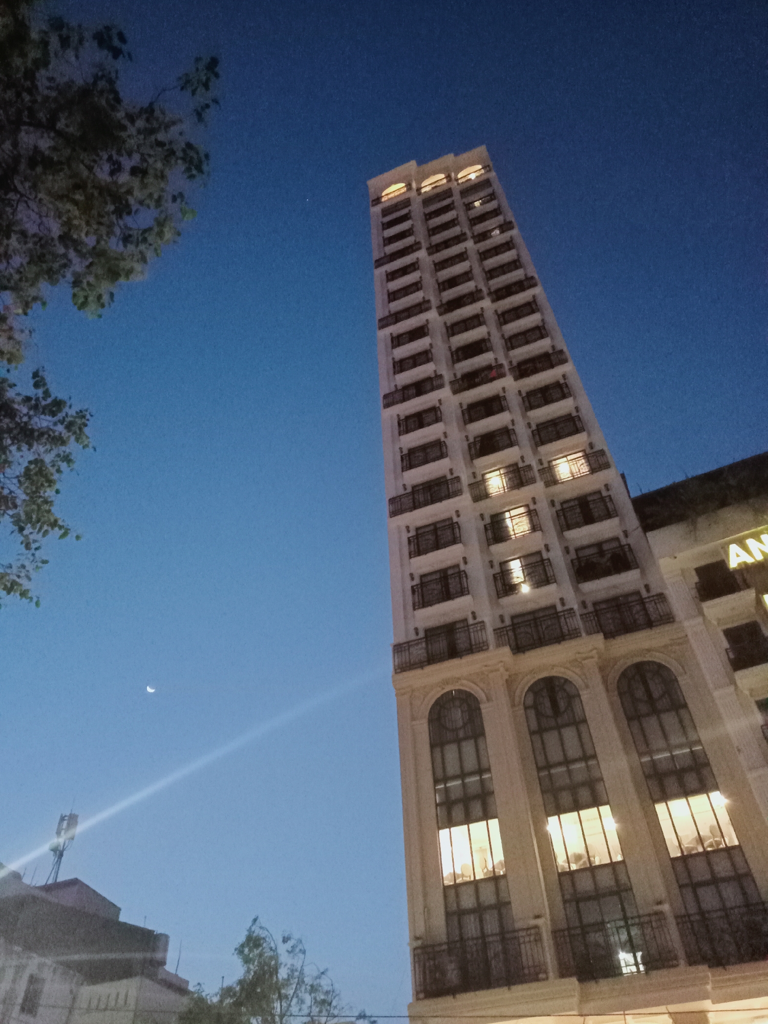 A tall, slender building dominates the right side of the frame, captured from a low, upward-looking perspective against a deep blue twilight sky. The building appears light-colored, possibly white or cream, and features numerous stacked balconies with dark railings on its upper floors. Many windows throughout the building glow with warm, yellow light, indicating internal illumination. The lower floors showcase larger, arched windows and more ornate architectural details.
To the left, branches of a tree with green foliage partially obscure the upper-left corner of the sky. A clear, waxing crescent moon is visible in the lower-middle left portion of the evening sky, accompanied by a faint, bright point of light (possibly a star or planet) higher up. A prominent, diagonal streak of bright light, likely a lens flare or a beam from a street light, cuts across the lower-left sky, originating near other partially visible buildings and an antenna structure.
The overall scene depicts an urban environment at dusk or early night, characterized by the clear sky and the artificial light emanating from the building. On an adjacent building visible on the far right, the partial letters "AN" can be seen as part of a sign. The setting is in Nha Trang, Vietnam.