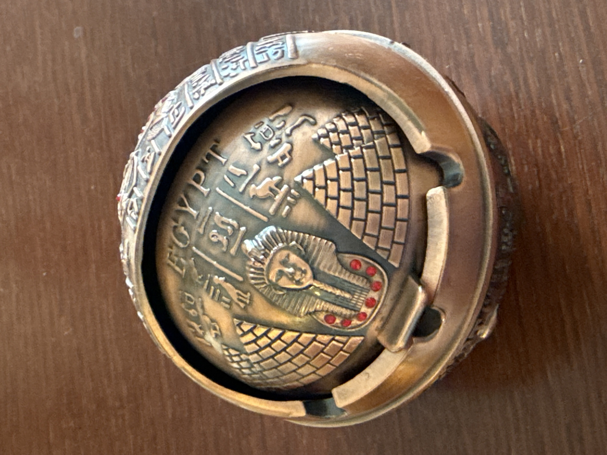 This is a close-up, top-down shot of a round, metallic object, possibly a ring or a pendant, with an Egyptian theme. The object is made of a bronze-colored metal and rests on a dark brown, wooden surface, likely a table.
The central feature of the object is a spherical element decorated with intricate carvings. This sphere appears to depict a sarcophagus with the face of a pharaoh, complete with a headdress. The sarcophagus is adorned with several small, red, jewel-like accents arranged in a circular pattern. Flanking the sarcophagus are representations of pyramids, textured with brick-like patterns. Encircling this central design are hieroglyphic symbols and the word "EGYPT" written in English in raised lettering. The outer edge of the object is a thick, metallic band, also with decorative carvings.
The lighting appears to be artificial, casting some shadows and highlighting the metallic sheen of the object. There are no people visible in the image. The setting is nondescript, focusing entirely on the artifact itself. The overall impression is one of an ornamental or collectible item related to ancient Egyptian culture.