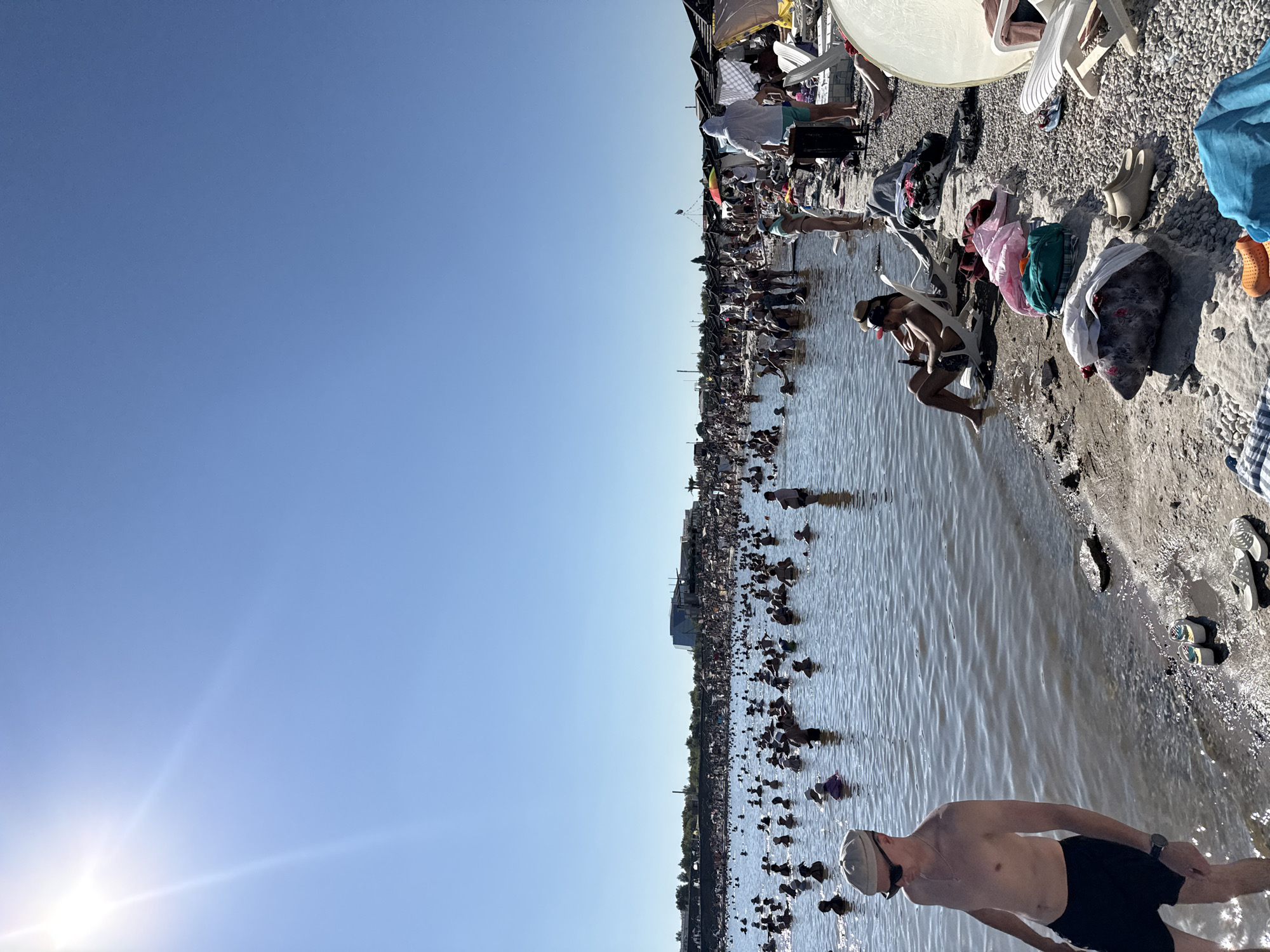 The image depicts a crowded scene at a salt lake in Sol-Iletsk, Russia, on a sunny day. Numerous people are in the shallow, murky water, some standing, others sitting. The water's edge is lined with a rocky, pebbly shore, where beach chairs, towels, and personal belongings are scattered. In the background, a dense crowd of people lines the shore, with some structures and trees visible beyond. The sky is clear and blue, with the bright sun in the upper left corner. A man in a cap and sunglasses is in the foreground on the left, facing away from the camera, standing at the water's edge.