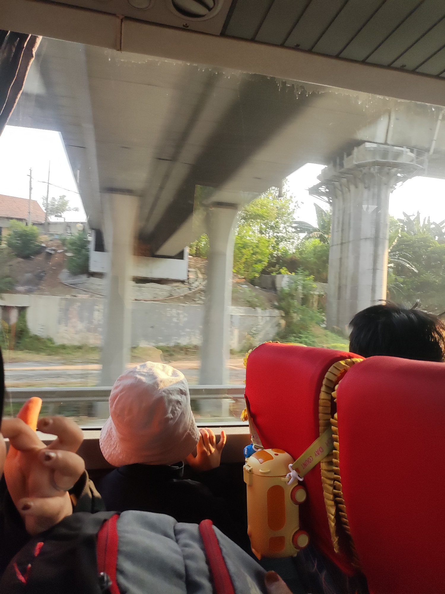 The image captures a view from inside a vehicle, likely a bus, looking out the window. In the foreground, two young children are visible from behind. One child, wearing a light-colored hat, is seated facing away from the camera. The other child's head and shoulder are visible to the right, with dark hair. In front of the child with the hat, a bright orange and yellow sippy cup with a cartoonish design is attached to the red seatback. A hand with painted fingernails is reaching out from the left side of the frame, towards the child with the hat.
Outside the window, a concrete overpass structure dominates the background, supported by large pillars. Beneath the overpass, there are glimpses of vegetation, including trees and bushes, and a building with a red roof. The lighting suggests it is daytime, possibly morning or late afternoon, with sunlight casting shadows. The overall scene appears to be a journey through a semi-urban or rural area, possibly on a road passing under an elevated structure. The presence of children and a sippy cup indicates a family or group outing. The location is identified as Sukaresmi, Indonesia, suggesting a setting within this region.