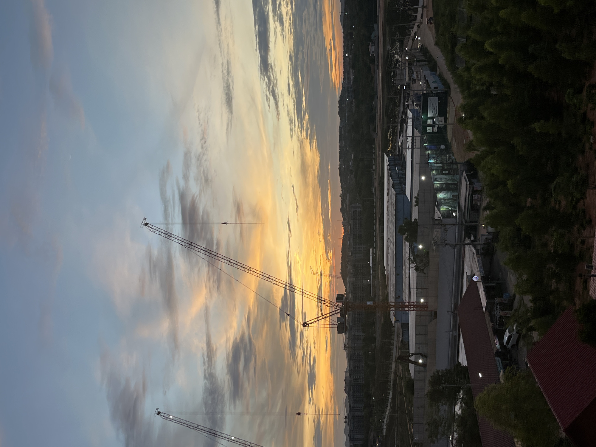 The image captures a twilight scene in Pattaya City, Thailand, featuring construction cranes against a sky streaked with orange and gray clouds. The cranes appear to be part of an ongoing construction project in the city. The location is likely an urban area, given the presence of buildings, trees, and infrastructure. Some people are on a walkway.