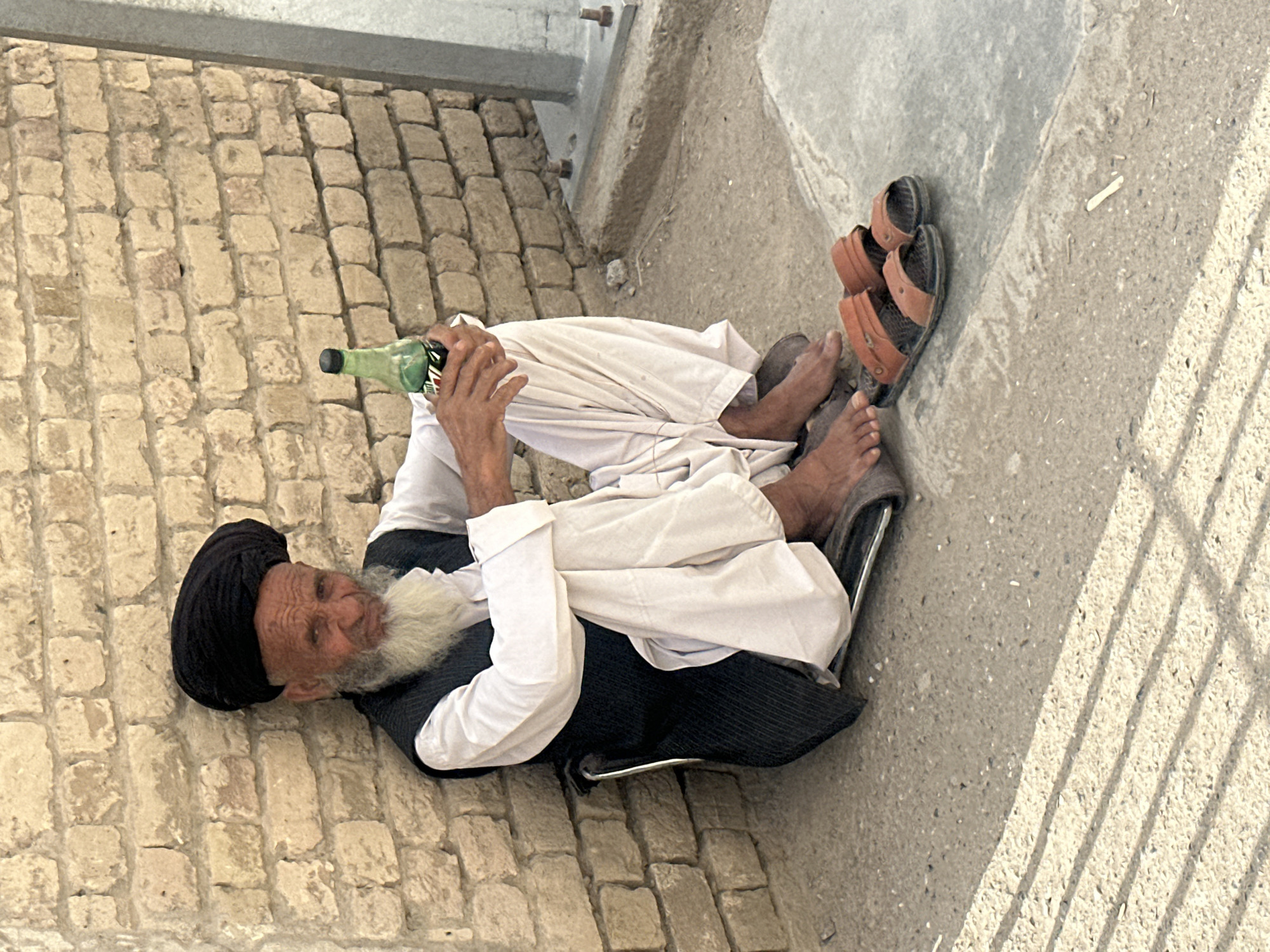 An elderly man wearing a black turban, white beard, white shirt and black vest is reclining in a metal chair next to a brick wall in Quetta City Tehsil, Pakistan. He is holding a green glass bottle in his left hand. His bare feet rest on a pair of brown sandals placed on the concrete floor. A metal beam is visible above. Shadows of a fence are cast on the ground.