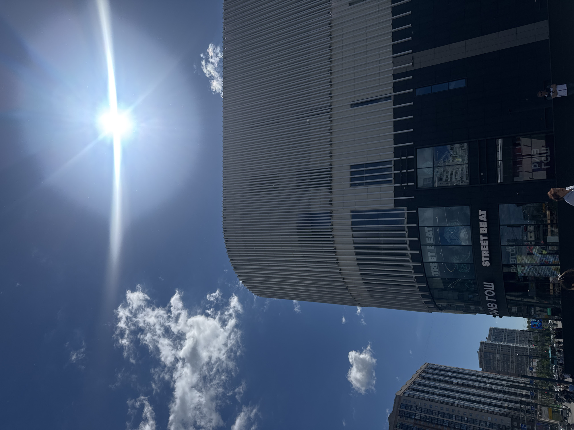 The image depicts a contemporary urban scene with a prominent, modern building. The building features a sleek facade with horizontal white louvers on the upper portion and dark glass panels on the lower levels. Through the glass, interior spaces or advertisements are visible, including text that reads "STREET BEAT" and "РИВ ГОШ". Several people are standing or walking near the building, some of whom are partially visible.
The sky is a bright, clear blue, with scattered white cumulus clouds. A strong sun is positioned in the upper left of the frame, casting a bright glare and lens flare across the image, suggesting it is midday. The sun's intensity indicates a sunny day.
In the background, other buildings, some appearing residential with multiple windows, are visible. The overall setting is a city street with elements of commercial or entertainment establishments, suggested by the "STREET BEAT" signage. The composition, with the camera angled upwards towards the building and sky, gives a sense of scale and grandeur to the architecture. The presence of people suggests the area is active and open to the public.