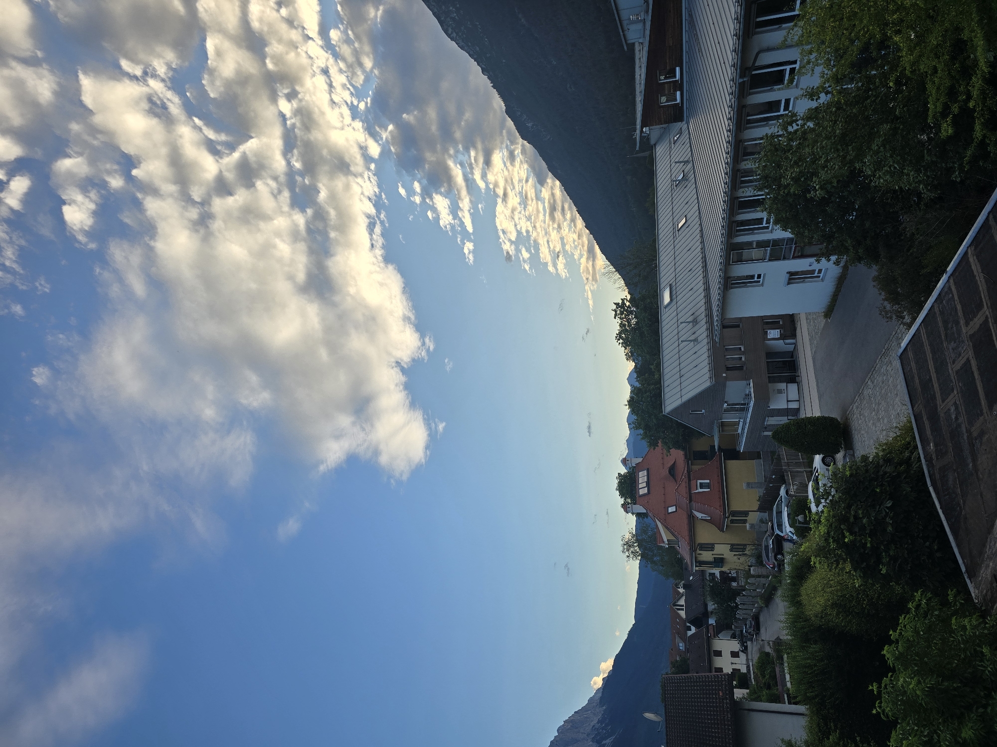 The image depicts a scenic view of Garmisch-Partenkirchen, Germany, bathed in the soft light of what appears to be late afternoon or early evening. The sky is a vibrant blue, punctuated by scattered white clouds, some of which are tinged with golden hues, suggesting the sun is beginning to set.
The foreground of the image is dominated by the upper floors of buildings with modern architectural styles, featuring large windows and metal siding. Adjacent to these buildings is a dense canopy of lush green trees, adding a natural element to the urban landscape. Further into the scene, a charming collection of houses with red-tiled roofs and yellow facades nestles against a backdrop of imposing, dark, and majestic mountains. These mountains, characteristic of the Bavarian Alps, rise sharply, with their peaks partially obscured by the hazy glow of the setting sun and some wispy clouds at their base.
A paved road winds through the residential area, with several parked cars visible, including a white car and a darker sedan. The overall atmosphere is one of tranquility and natural beauty, seamlessly integrated with the human settlement. The orientation of the image is sideways, presenting the scene from a slightly elevated perspective, as if captured from a balcony or window. No people are discernible in the frame, and there is no visible text.