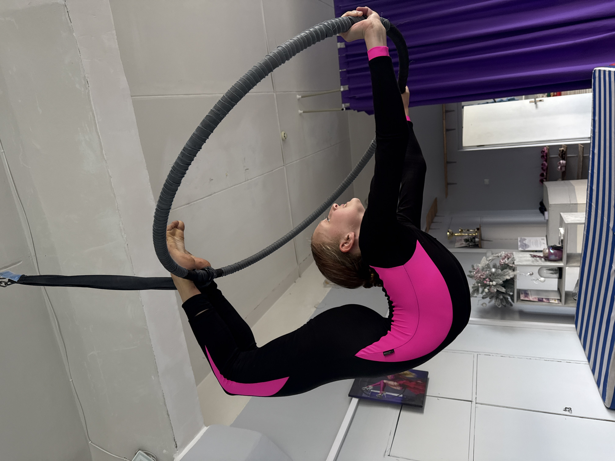 In Dnipro, Ukraine, a young female gymnast is captured mid-performance on an aerial hoop. She is wearing a black and neon pink leotard, her body contorted in a challenging pose as she grips the hoop. The hoop itself is suspended from the ceiling by a black fabric strap. The background reveals a studio setting with white walls and ceiling, along with purple curtains and shelves filled with decorative items and trophies, suggesting a dedicated space for training and performance. The lighting appears to be artificial, casting a bright glow on the scene.