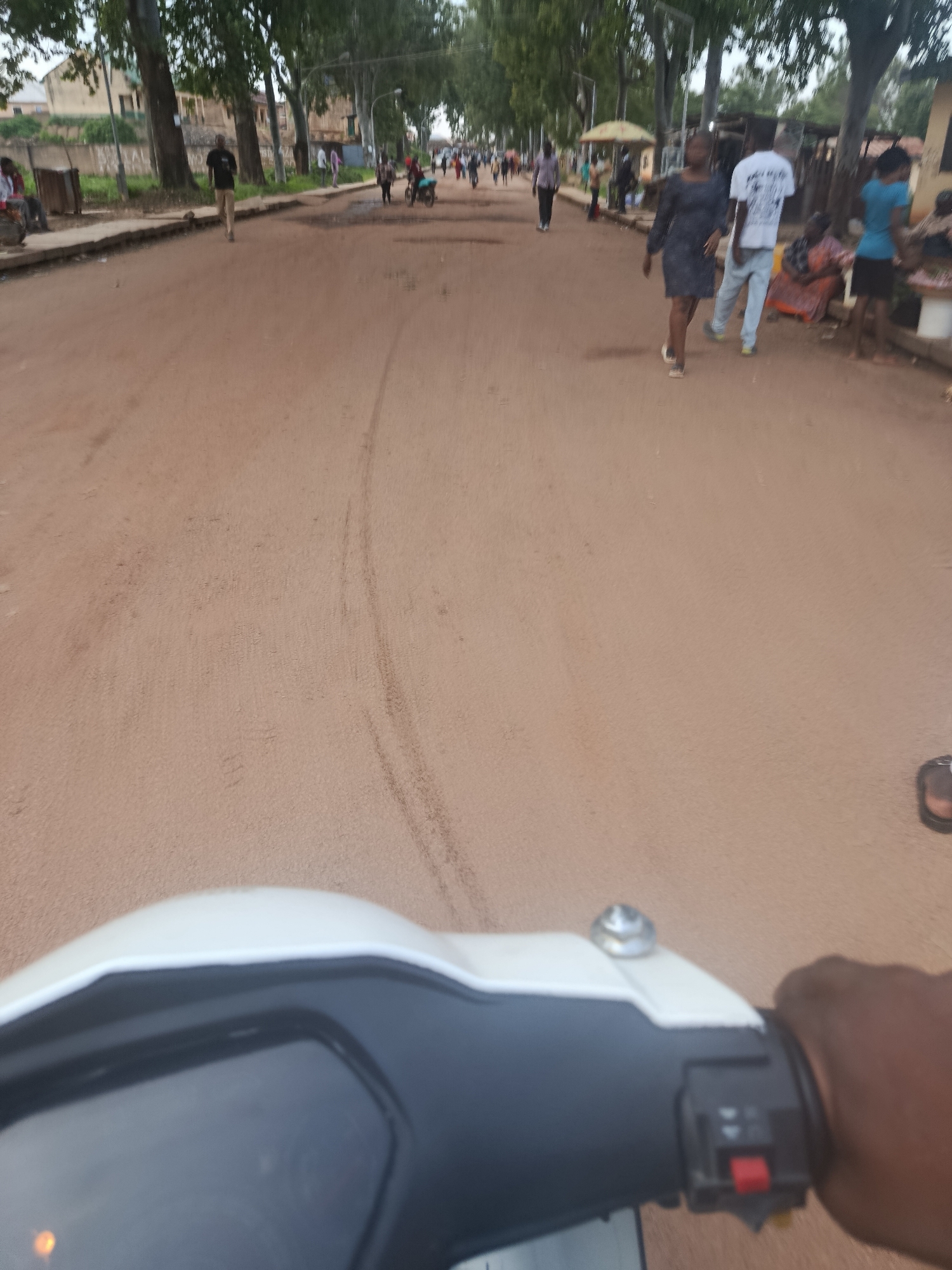 The image depicts a street scene in Rafin Par, Nigeria, characterized by a dirt road and a line of trees and buildings. The perspective is from a person riding a scooter, with the front of the scooter visible in the foreground. Several individuals are present on the street, walking and standing. Some are further down the road, appearing to engage in various activities, while others are closer, with a woman in a dark dress and a man in a white t-shirt and light-colored pants being more prominent. The environment suggests a typical urban or semi-urban setting in Nigeria, with a mix of residential or commercial structures lining the street. The road surface is unpaved, indicating a less developed area. The lighting suggests it is daytime, with the sky being overcast.