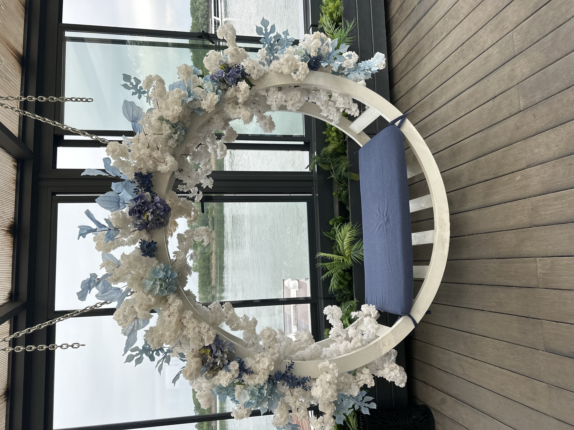 A white, circular swing chair hangs suspended by metal chains in a building with floor-to-ceiling windows. The chair is adorned with white and blue artificial flowers and a blue cushion. The flooring appears to be wooden planks. Through the windows, water and a distant shoreline are visible. The building is possibly located in Vidin, Bulgaria.