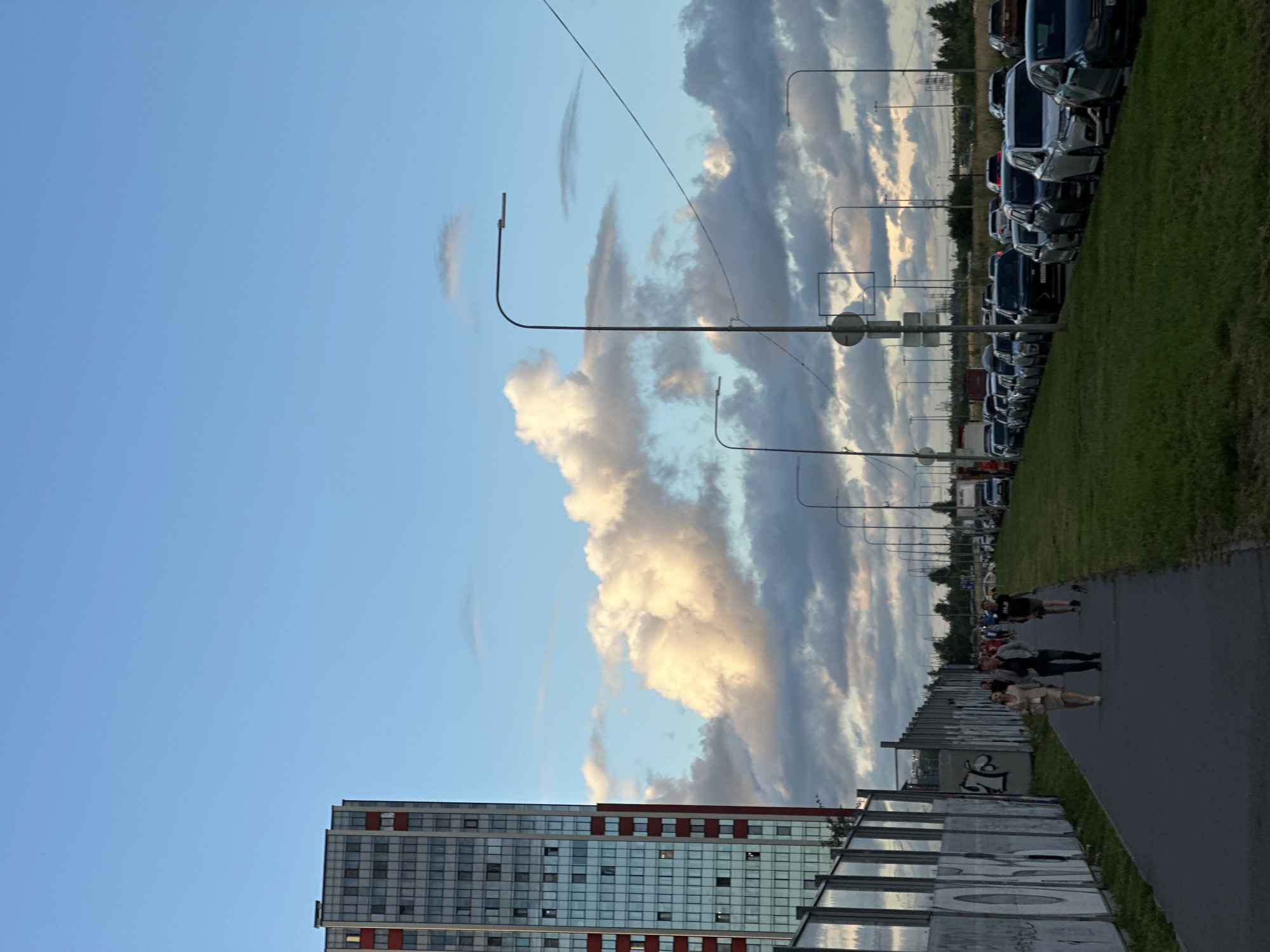 The image depicts a scene on a sunny day in Kudrovo, Russia, with a clear blue sky and large white clouds. A tall, modern apartment building with red accents on its facade dominates the left side of the frame. In the foreground, a paved pedestrian walkway runs alongside a grassy area and a long fence covered in graffiti. Several people are walking on the walkway. To the right of the walkway, a line of parked cars stretches into the distance along a road. Streetlights are visible along the road. The overall impression is an urban setting with residential buildings, infrastructure, and signs of daily life.