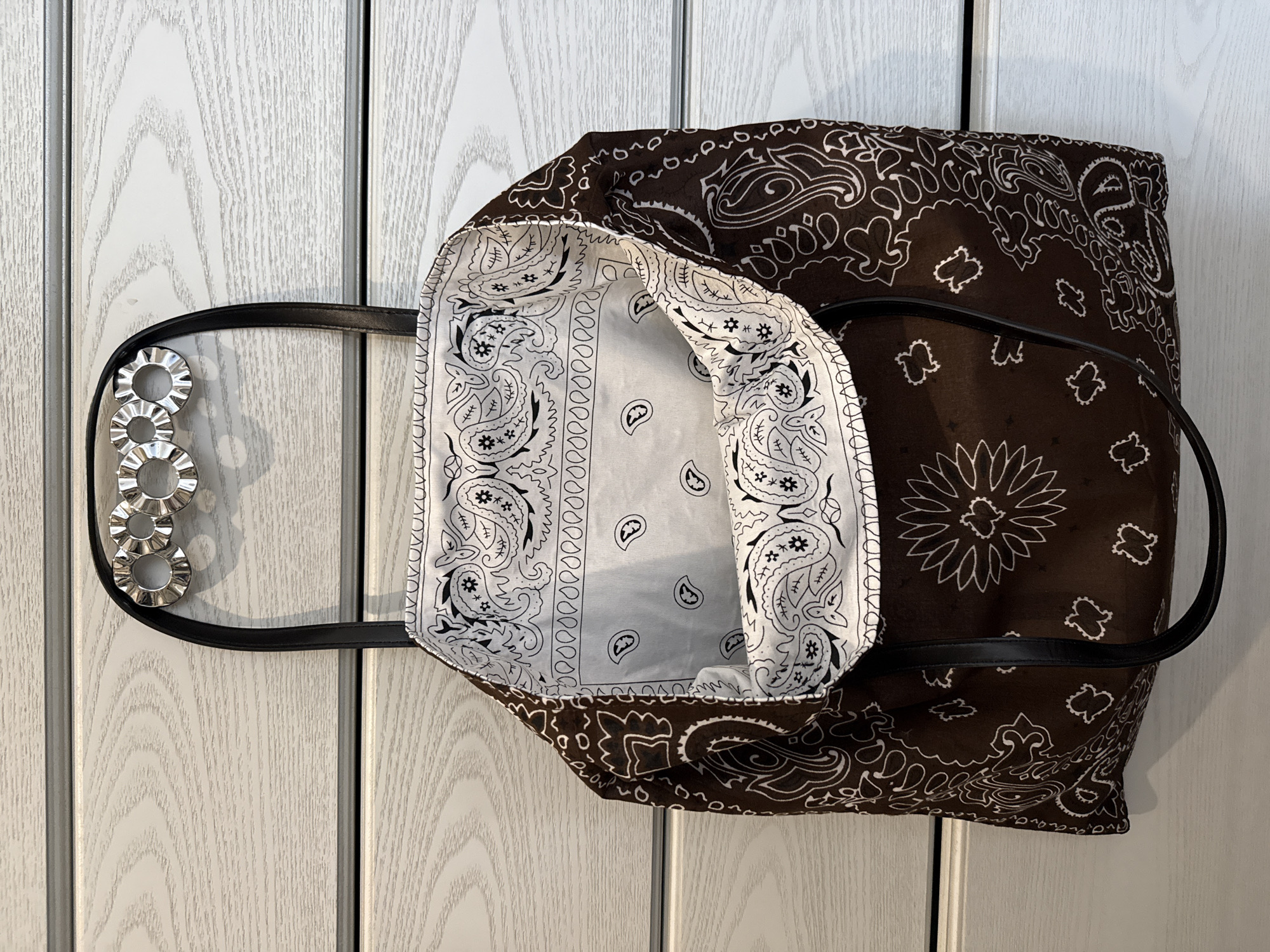 A brown and white bandana-patterned tote bag with black handles hangs on a white wood paneled closet door. The tote bag's interior is visible and lined with white bandana-patterned fabric. There are four silver metal rings attached to one of the handles. The location in the city of Votkinsk, Russia is not discernible from the image.