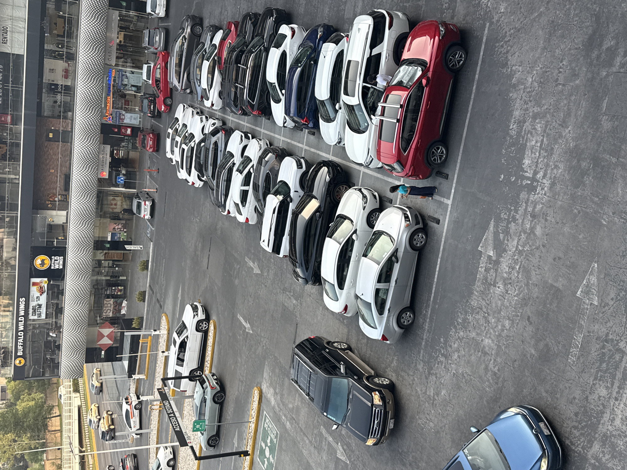 The image shows an aerial view of a parking lot adjacent to a building in San Bernardino Tlaxcalancingo, Mexico. The parking lot is filled with several rows of parked cars, primarily in shades of white, black, blue, and red. Most cars appear to be newer models. Arrows painted on the ground indicate traffic flow. A person in a blue shirt is standing next to a red car.
The building next to the parking lot houses multiple businesses, including a Buffalo Wild Wings restaurant. Signs for other businesses, such as "Farmacias Especializadas" (Specialized Pharmacies) and a brand labeled "GABIN," are visible.
The presence of a meeting point labeled "PUNTO DE REUNIÓN" and signs indicating "ENTRADA" (Entrance) and "SALIDA" (Exit) suggest this is a commercial area.