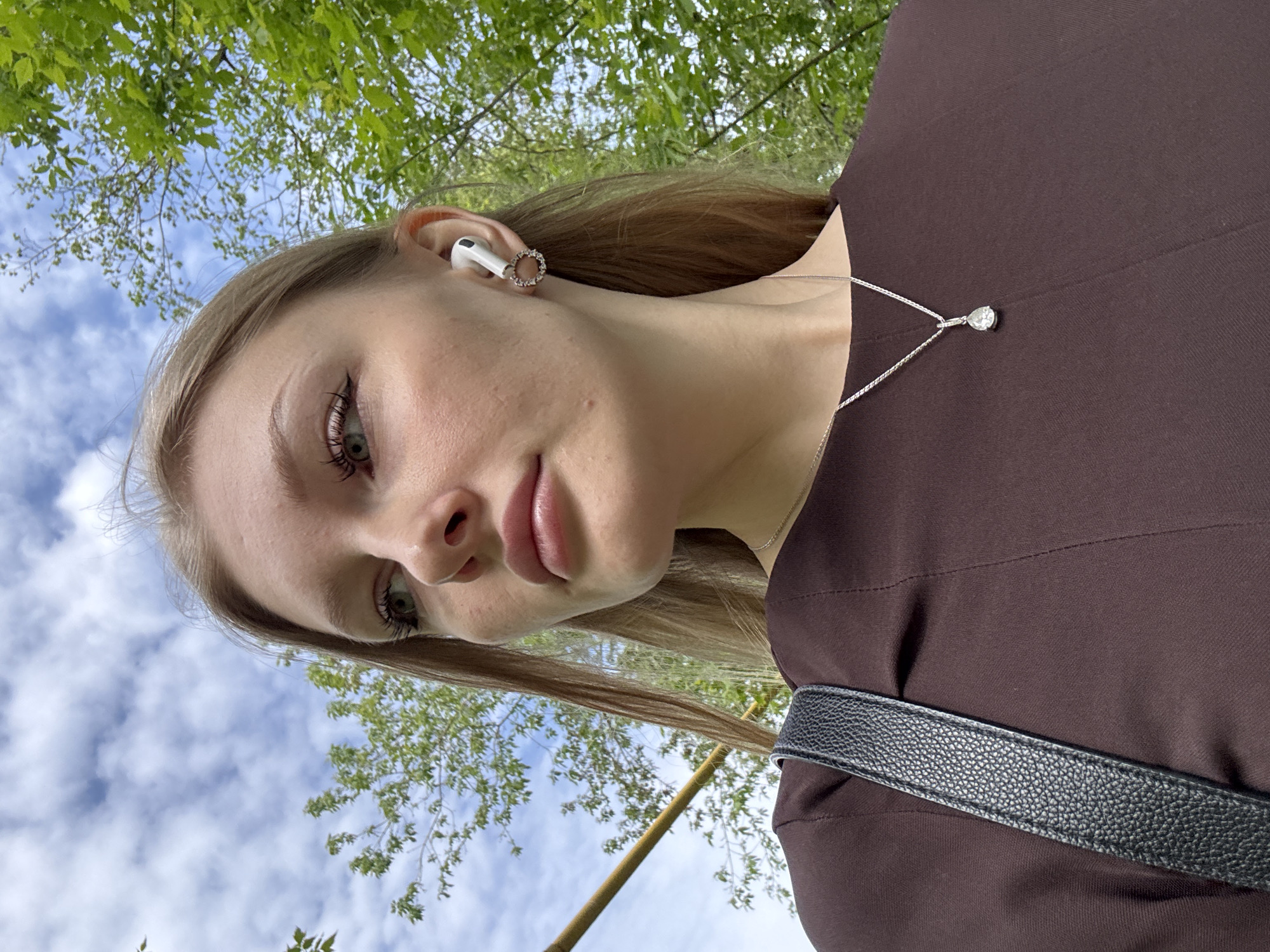 A low-angle, close-up shot of a young woman in Perm, Russia. The woman is fair-skinned with light brown hair, wearing an AirPod in her left ear, adorned with a ring earring. She wears a brown dress and a silver necklace with a teardrop pendant. A black purse strap is visible across her chest. The background shows blue sky with clouds, green trees, and a bamboo branch.