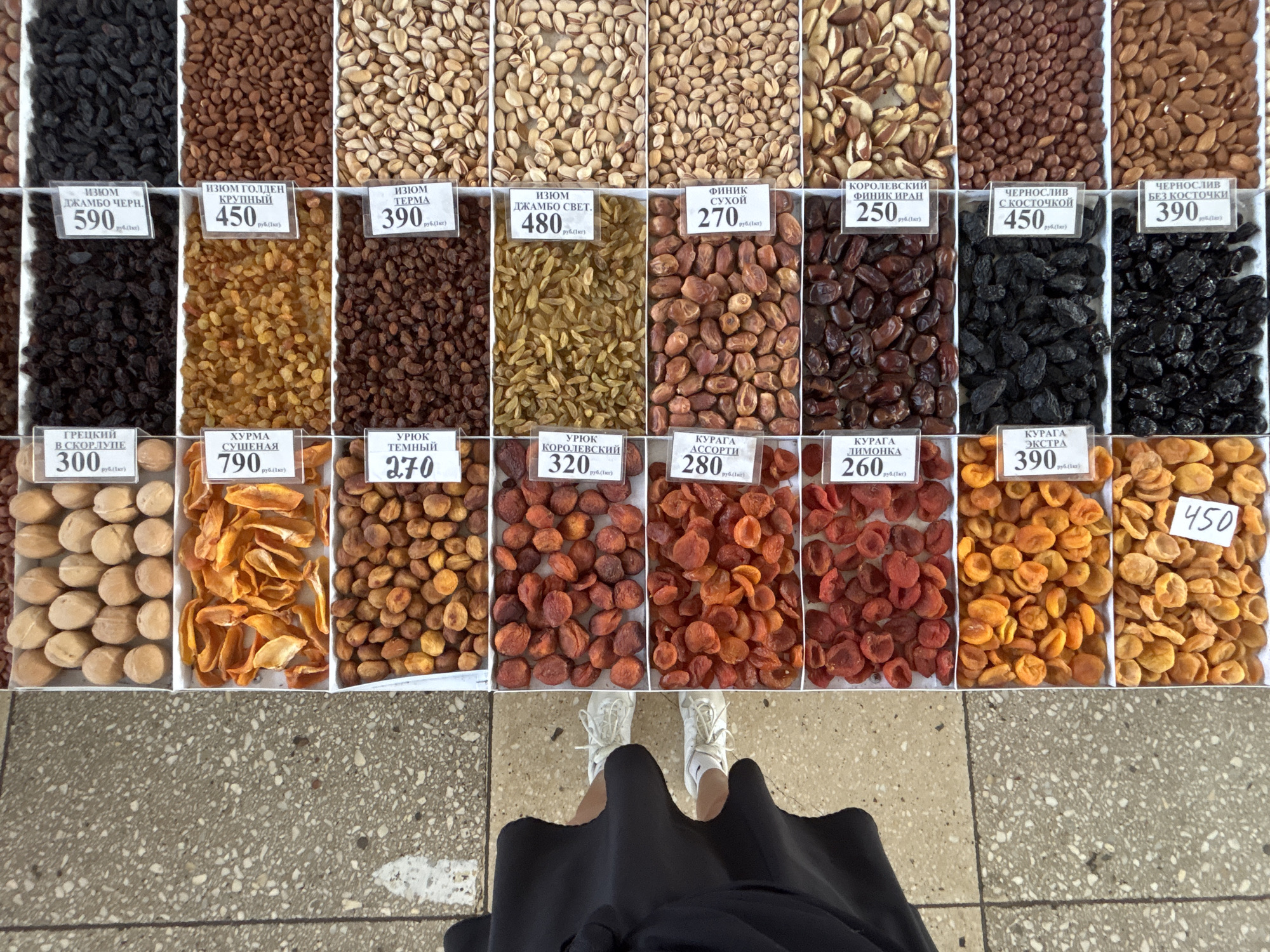 The image shows a display of dried fruits and nuts at a market, most likely in the city of Chaikovsky, Russia. There are several varieties including raisins, dried apricots (kuraga and uryuk), dates (finik), prunes (chernosliv), and walnuts, neatly arranged in separate sections or trays. Each section has a small label with the name of the product in Russian and its price per kilogram. A person wearing a black skirt and white sneakers is standing in front of the display. The floor is tiled in a speckled gray and white pattern.