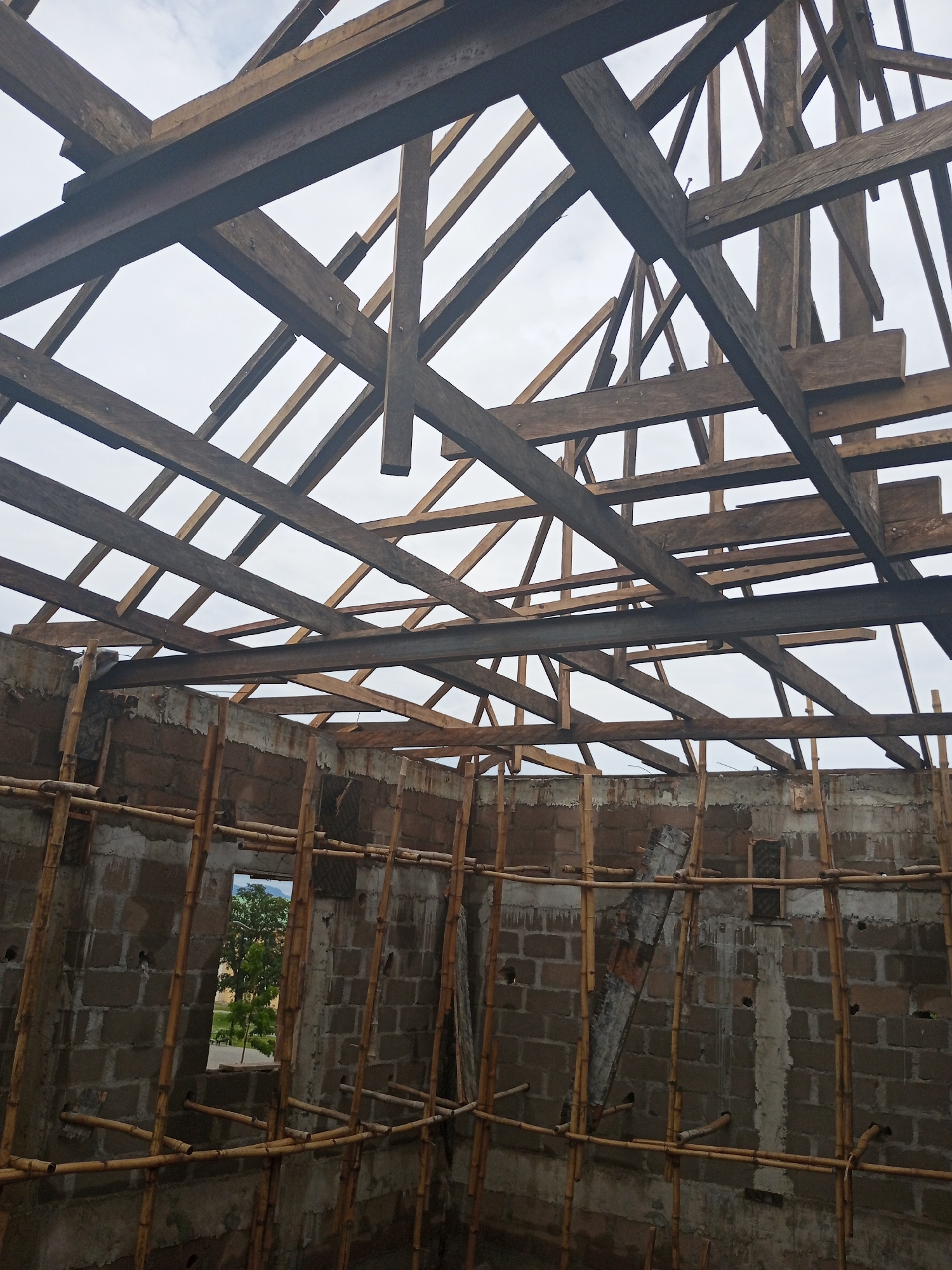 The image depicts the interior of a building under construction in Jalingo, Nigeria. The structure features cinder block walls and a complex wooden roof truss system, with a steel beam integrated into the framework. Bamboo scaffolding is visible, supporting the construction work. Through an opening in the wall, a tree and distant landscape are visible, suggesting an outdoor environment beyond the immediate construction site. The sky is overcast. There are no people or animals present in the scene.