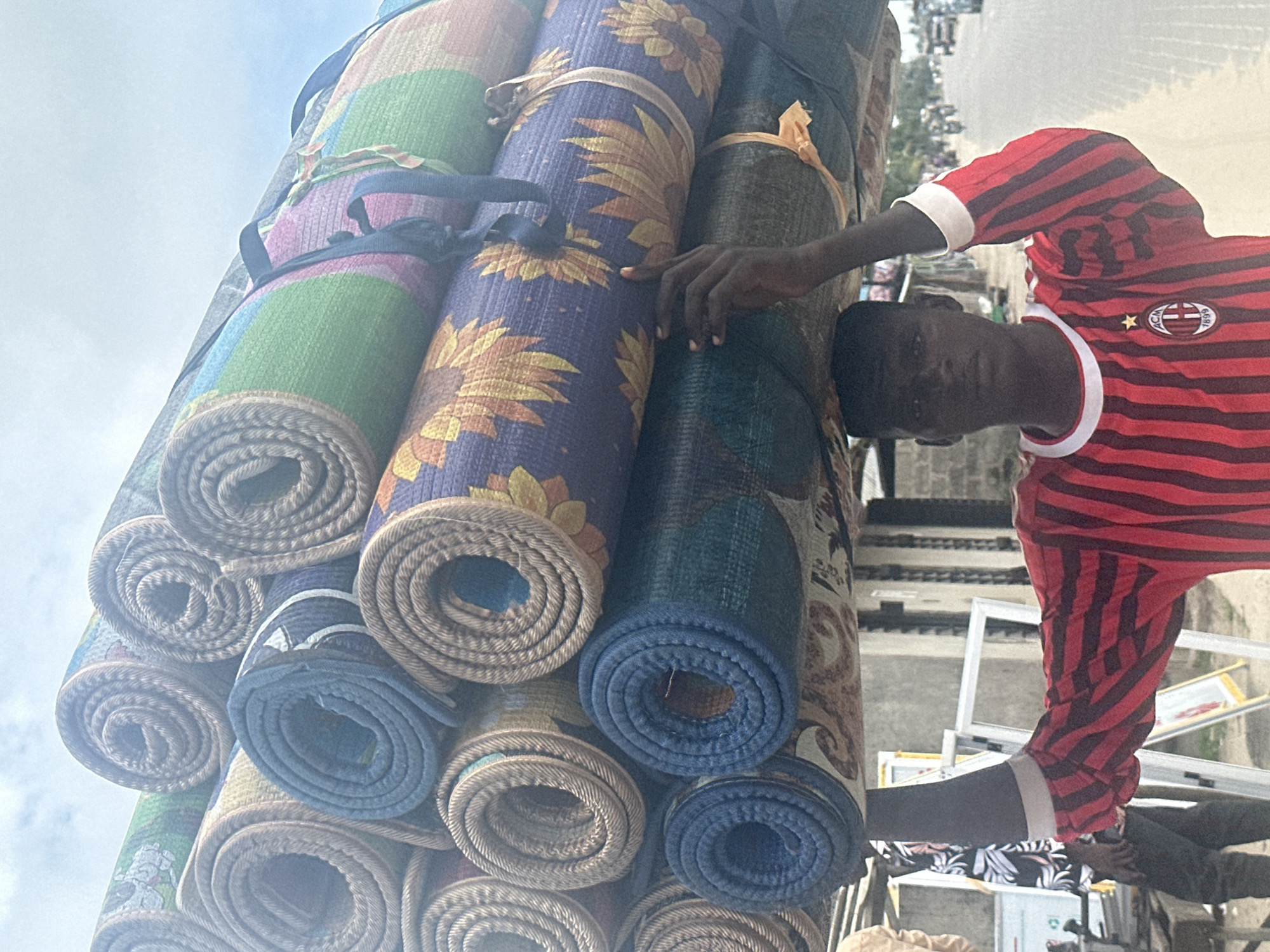 A young Black male is shown from the chest up, carrying a large stack of rolled-up rugs on his head. He is wearing a red and black striped shirt with a white collar and a logo on the left chest. The rugs are colorful and feature various patterns, including sunflowers and geometric designs. The background depicts a street scene in Badagry, Nigeria, with buildings and other people visible. Some of the buildings appear to be shops or market stalls. Vehicles, including motorcycles and cars, are present on the road. The sky is overcast. The image seems to capture a moment of daily life and commerce in the city.