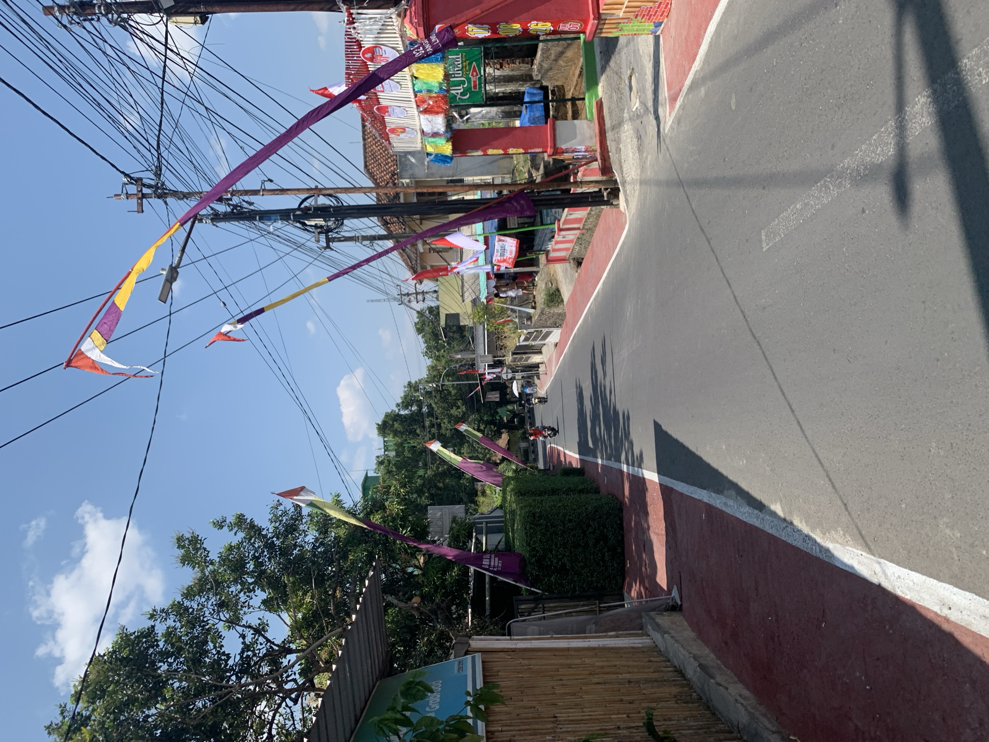 The image shows a street in Ciamis, Indonesia, decorated with numerous red and white Indonesian flags, as well as purple and yellow pennants. The flags are attached to poles and strung across the street, suggesting a festive occasion, possibly Independence Day celebrations. The street itself is asphalt, with a red-painted pedestrian walkway on the left. On the right side of the street, there are buildings, some with red and white decorations, and a red archway with colorful banners. Several utility poles with a dense network of wires are visible throughout the scene. A few people are present; two individuals are riding motorcycles in the distance, and a person is standing near a stall on the right. The sky is clear and blue with a few clouds. The overall impression is a vibrant street scene in a populated area.