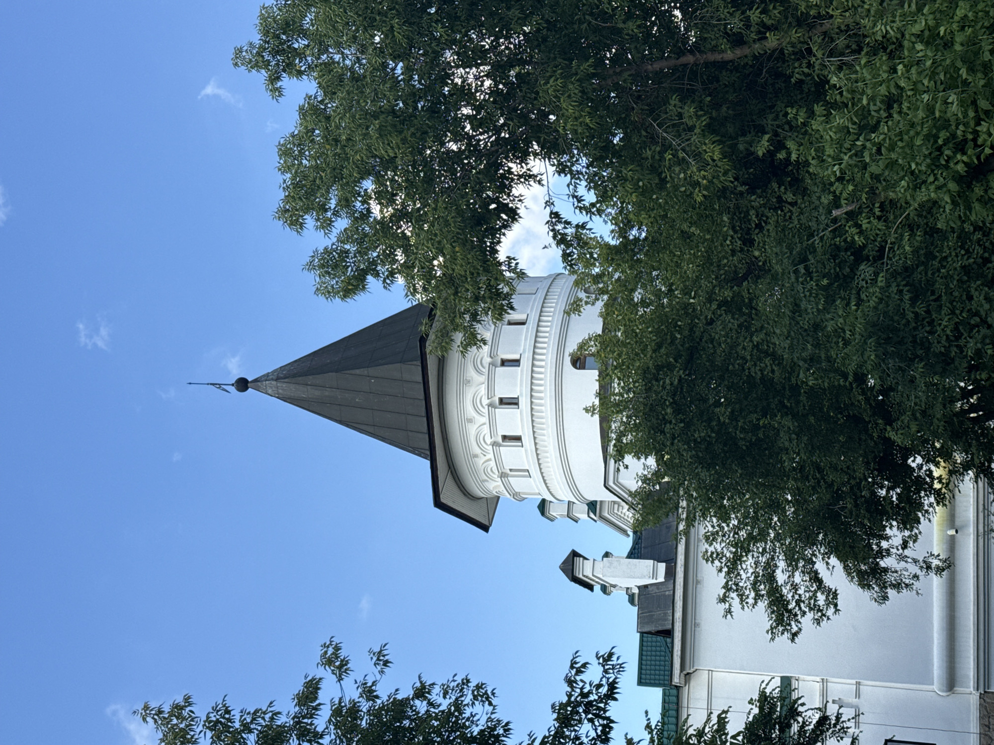 The image shows a white tower with a dark, conical roof, peeking through lush green trees. The tower has decorative circular elements and arched windows. It appears to be part of a larger building, with hints of other white structures and a green roof visible. The sky above is a clear blue with a few wispy clouds. The overall impression is one of a historical or architectural landmark, possibly in a park or garden setting. This is the Church of the Holy Trinity at Ganina Yama, a significant religious site located near Yekaterinburg, Russia.