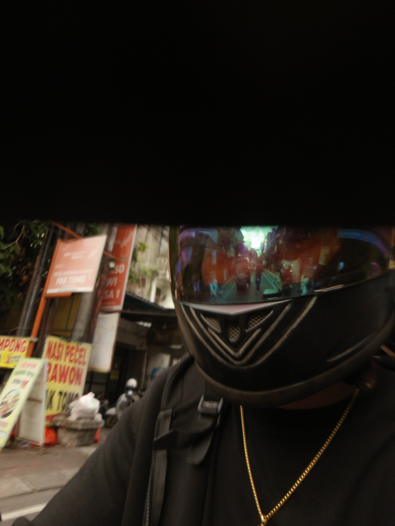 The image shows a first-person perspective of someone wearing a black helmet and a black jacket, with a gold chain visible around their neck. The helmet's visor reflects a street scene with buildings, lights, and other vehicles, suggesting the person is on a motorcycle or scooter. In the background, there are signs in Indonesian, with some text legible, such as "MPONG", "NASI PECEL", and "RAWON", indicating a food stall or restaurant. Another sign reads "PAK TOMO". The context of these signs and the overall urban environment suggests the location is within the city of Denpasar, Indonesia. A person wearing a white helmet is visible on a motorcycle in the middle ground. The street appears to be busy with traffic and pedestrians.