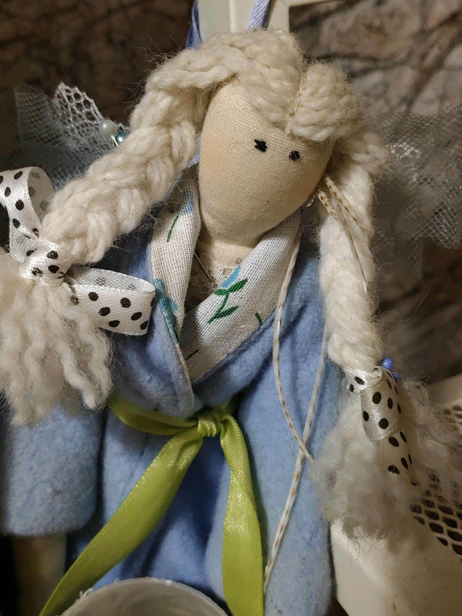 A close-up, medium shot captures a handmade fabric doll with blonde yarn hair in braids. The doll wears a light blue fabric coat, a white fabric scarf with a floral pattern, and is adorned with black and white polka dot ribbons. A bright green ribbon is tied around the doll's waist. The background consists of a blurred brown and gray textured surface. The image composition is tight, focusing on the doll's upper body and facial features, which are simple black stitches for eyes and a mouth. The doll's left side is more in focus than the right. The doll is located on display in Rostov-on-Don, Russia.