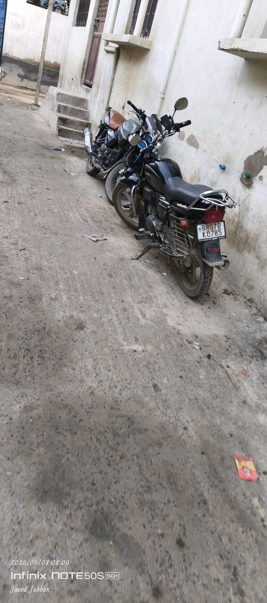 Two motorcycles are parked side-by-side on a dirt and concrete path in front of a light-colored building. The motorcycle in the foreground is black with a brown seat and a license plate that reads "BR07B K0785". The other motorcycle is partially visible behind it. The building has a dark brown door and a set of concrete steps leading up to it. The path is littered with small debris and a few colorful wrappers. The overall scene appears to be in an urban or semi-urban environment, possibly in Bahera, India. The image is taken from a low angle, looking down at the motorcycles and the path.