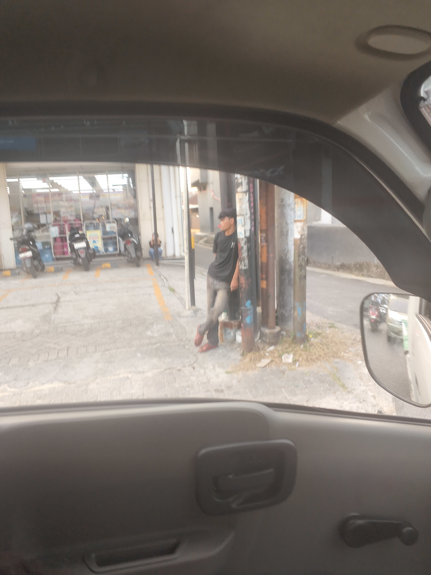 The image depicts an outdoor scene, possibly near a convenience store or gas station, on a paved area with yellow markings. A young man wearing a black t-shirt and grey pants, with red shoes, is leaning against a pole. He is looking to his left and his right hand is resting on the pole. In the background, several motorbikes are parked. To the right, a side-view mirror of a vehicle shows traffic, including what appears to be a white car and another vehicle with its lights on. The overall environment is slightly blurry, suggesting the photo was taken from inside a moving vehicle. The presence of motorbikes and the style of the buildings and street furniture are consistent with urban settings in Indonesia. Specifically, the scene is likely located in the city of Bandar Lampung, Indonesia, given the characteristic features observed.