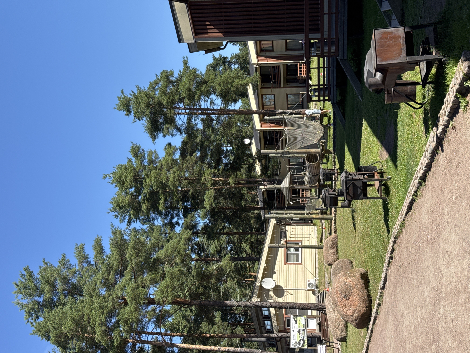 The image depicts a clear, sunny day in what appears to be a resort or recreational area in Ozerki (Krasnodolinskaya), Russia. Dominating the scene are tall, mature pine trees set against a bright blue sky. In the foreground, a gravel pathway curves along the edge of a grassy area bordered by small stones. The grassy area features several large, rounded boulders and multiple metal barbecue grills. Behind the grassy area, a cluster of light-colored, wooden buildings with brown trim and balconies are visible. A children's play structure with netting and wooden elements is situated between the buildings and the foreground. Two people, appearing to be children or young adults, are visible near the play structure. The overall environment suggests an outdoor leisure facility with accommodations.