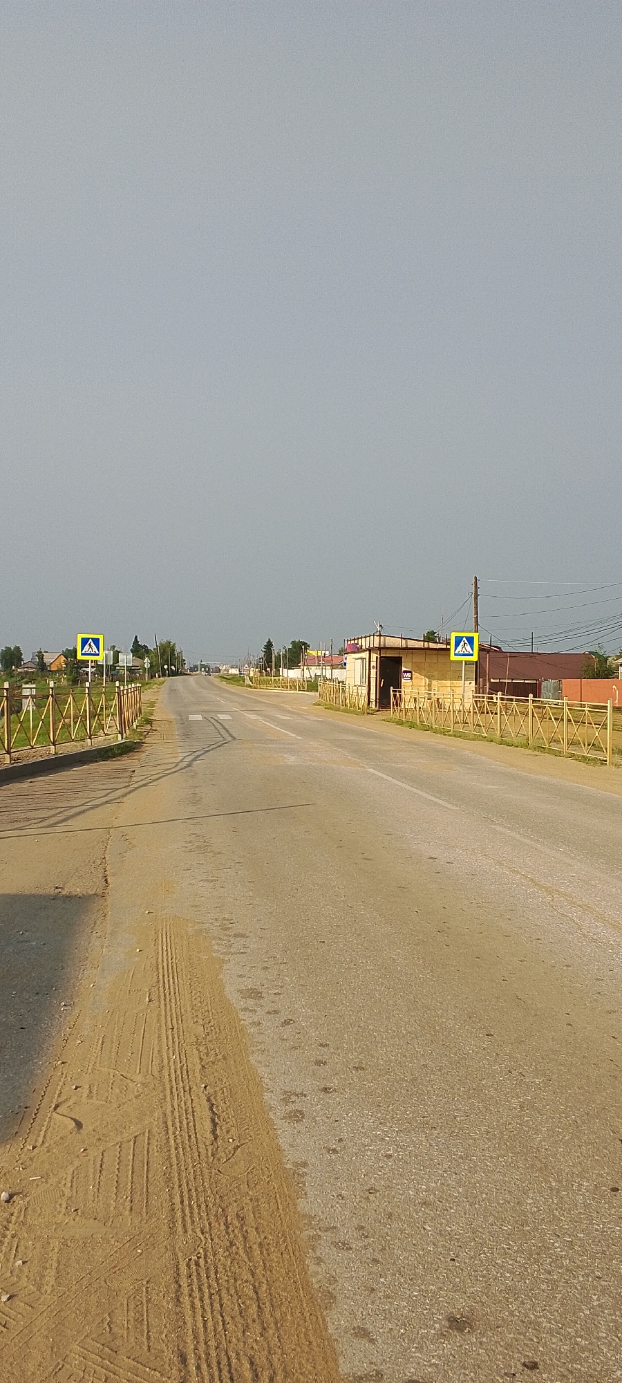 The image depicts a two-lane asphalt road stretching into the distance under a hazy, overcast sky. The road has a white line dividing the lanes and appears to be in a rural or semi-rural setting, indicated by the presence of houses and vegetation on either side. On the left side of the road, a pedestrian crossing sign is visible, a bright yellow square with a black silhouette of a person walking. A low, yellow fence runs along the roadside. On the right side, a similar yellow fence borders the road, and further down, a small, light-colored building with a dark entrance and a sign reading "WB" is situated. The ground on the left side of the road is covered in sand, with visible tire tracks. The overall impression is a quiet, somewhat desolate road leading through a settlement, likely in Tulagino, Russia, given the context provided. There are no people visible in the image.