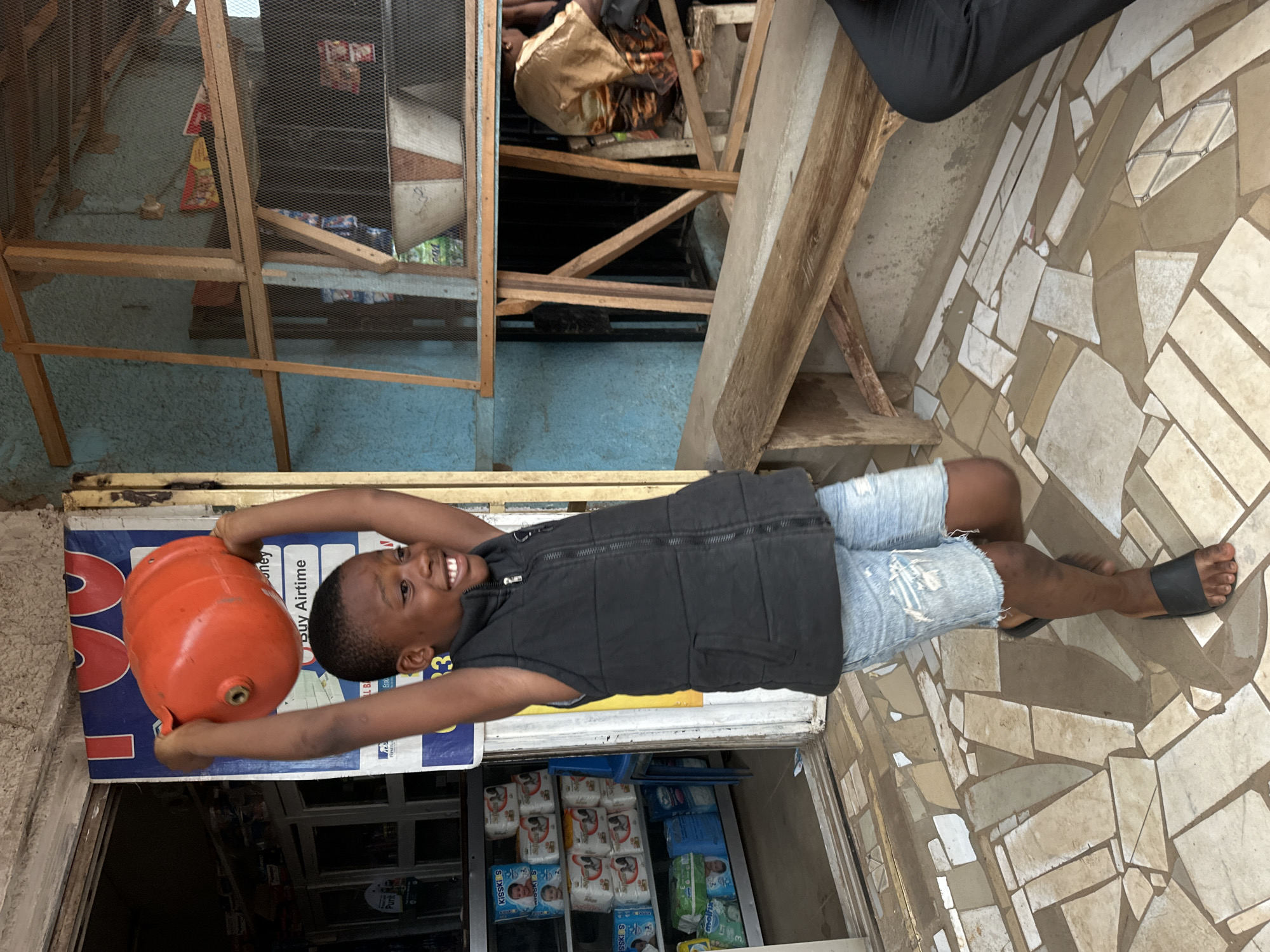In Narayi, Nigeria, a young boy, possibly around 8-10 years old, stands outside a shop. He is wearing a black vest over a blue shirt and distressed denim shorts, with black flip-flops on his feet. The boy is holding an orange propane tank above his head with both hands, his arms raised in a triumphant or playful gesture. He has a wide, joyful smile and appears to be mid-stride, with his right leg forward and his left leg bent.
Behind the boy, a shop entrance is visible, revealing shelves stocked with packaged goods, including diapers and what appear to be other household items. A blue advertising poster with red lettering is tacked to the wall near the entrance. To the right of the shop entrance, a wooden structure with shelves, covered by a mesh material, displays more items. In the background, a woman is seated on a stool, facing away from the camera, partially obscured by the wooden structure. The ground is paved with irregular-shaped tiles in various shades of beige and white. The overall scene depicts a lively street-level commercial area.